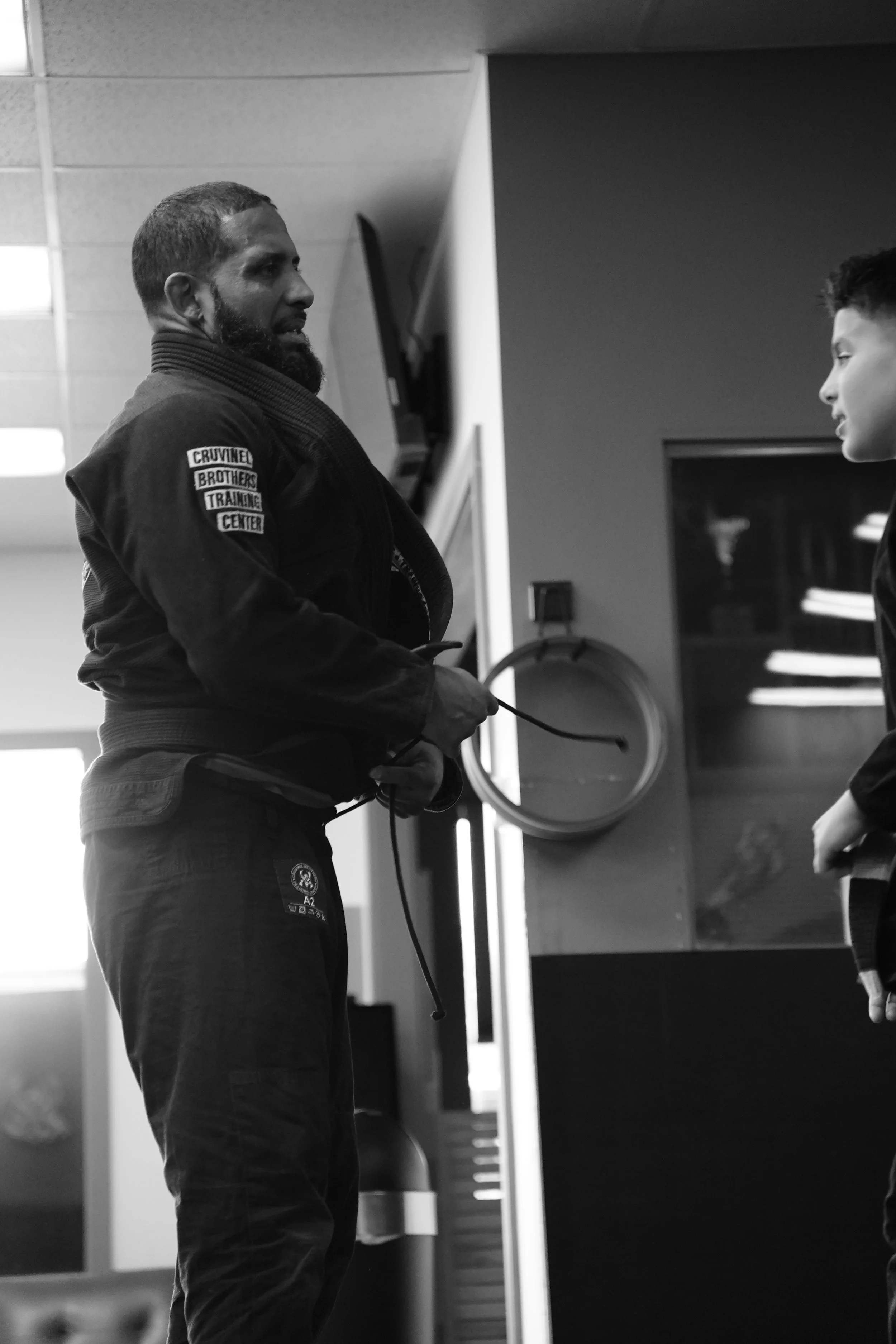 A man with a beard wearing a martial arts gi and holding a belt is looking at a young person in a training facility.