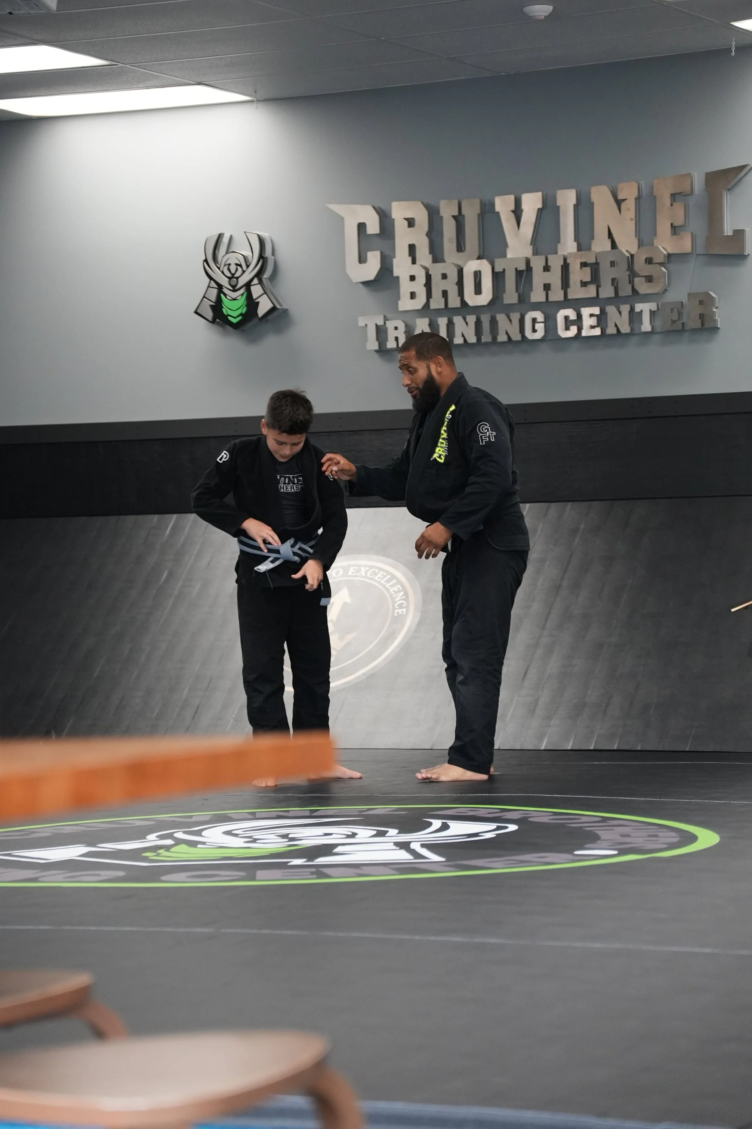 A martial arts instructor and student in black uniforms training in a gym at Cruzline Brothers Training Center. The instructor is helping the student with a gi tie, and they are barefoot on a mat with a logo.