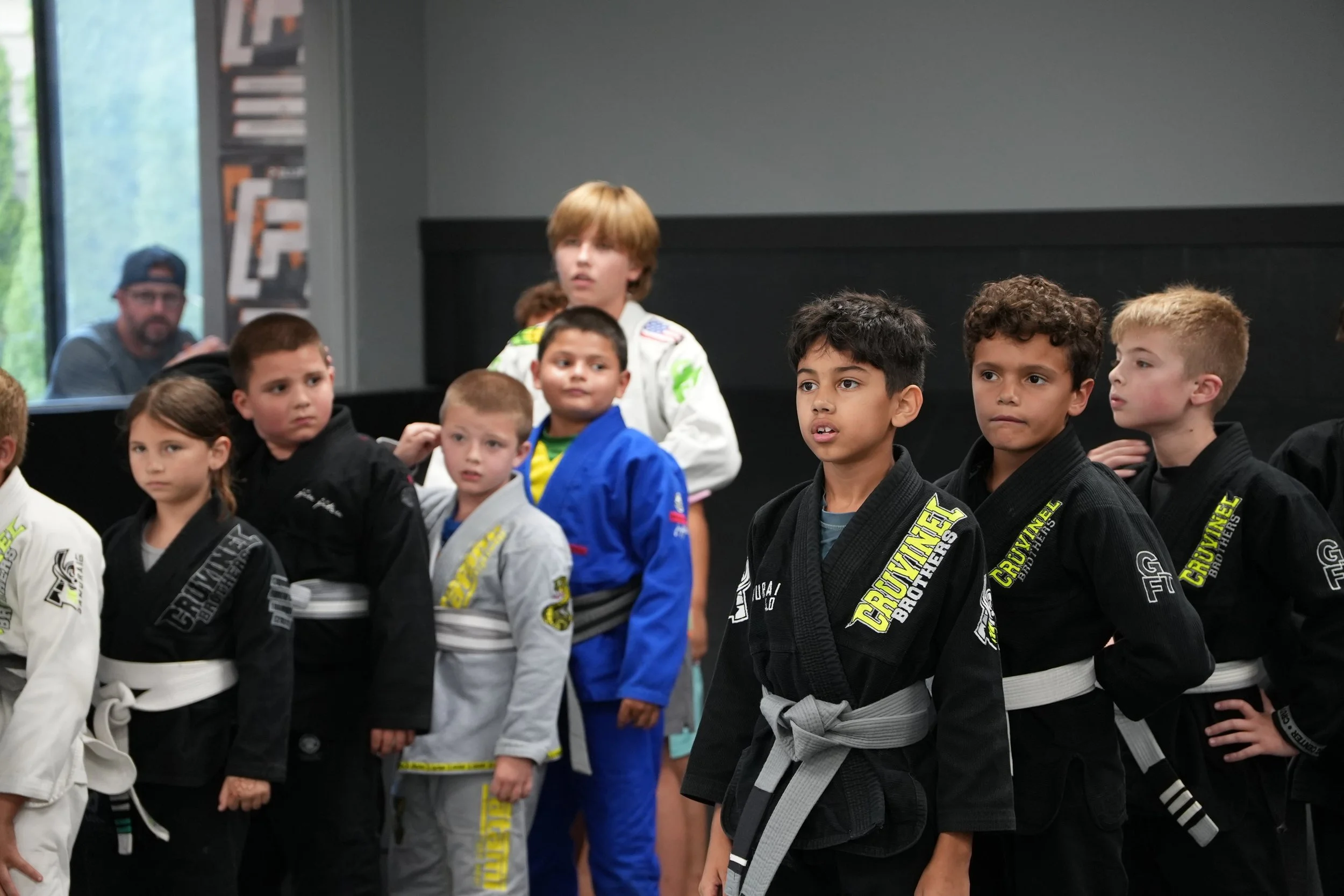 A group of children dressed in Brazilian Jiu-Jitsu uniforms, lined up indoors, with a man observing through a window in the background.