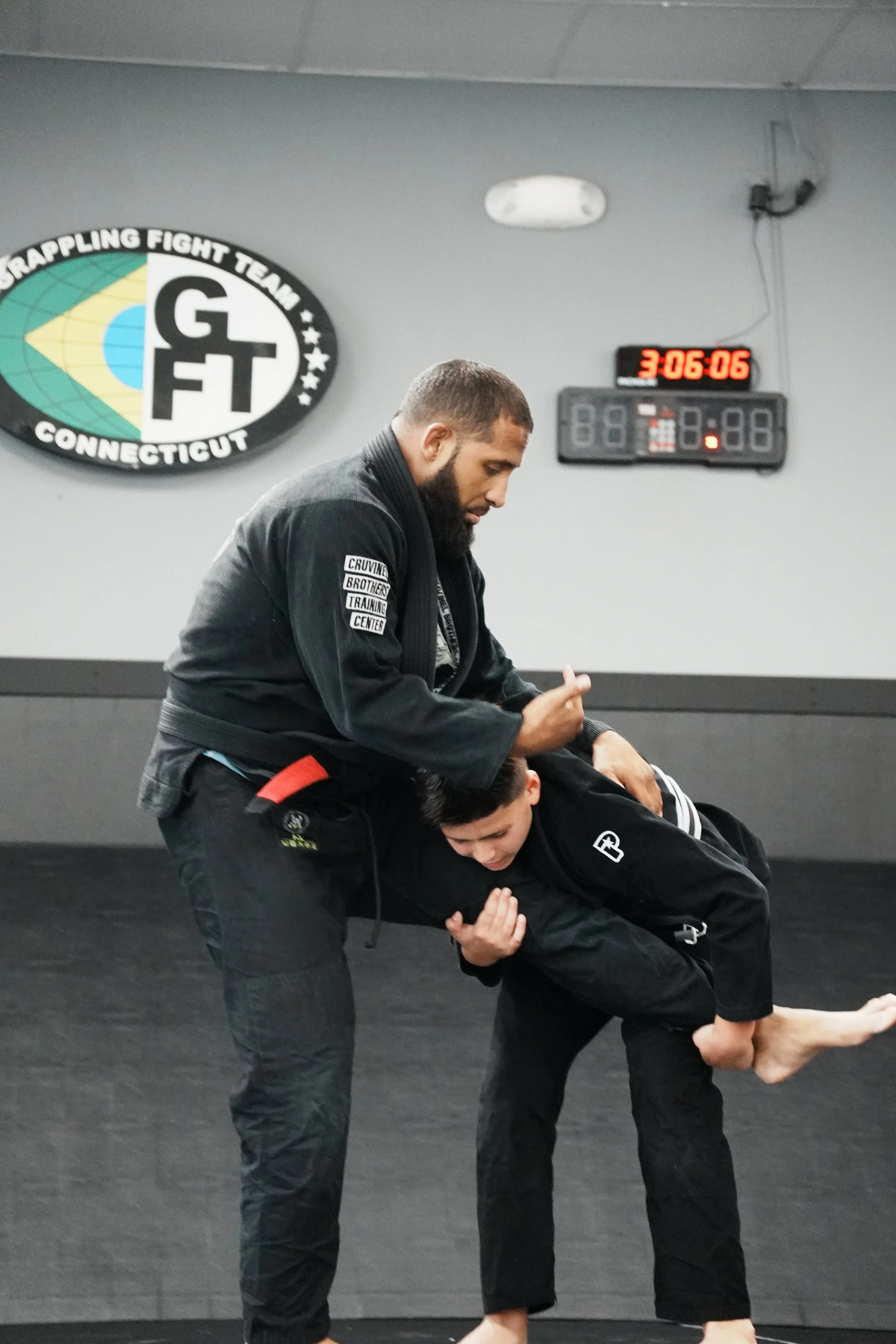 Two martial artists practicing Brazilian Jiu-Jitsu in a gym, one standing and the other on the ground, with a digital clock showing 3:06:06 in the background.