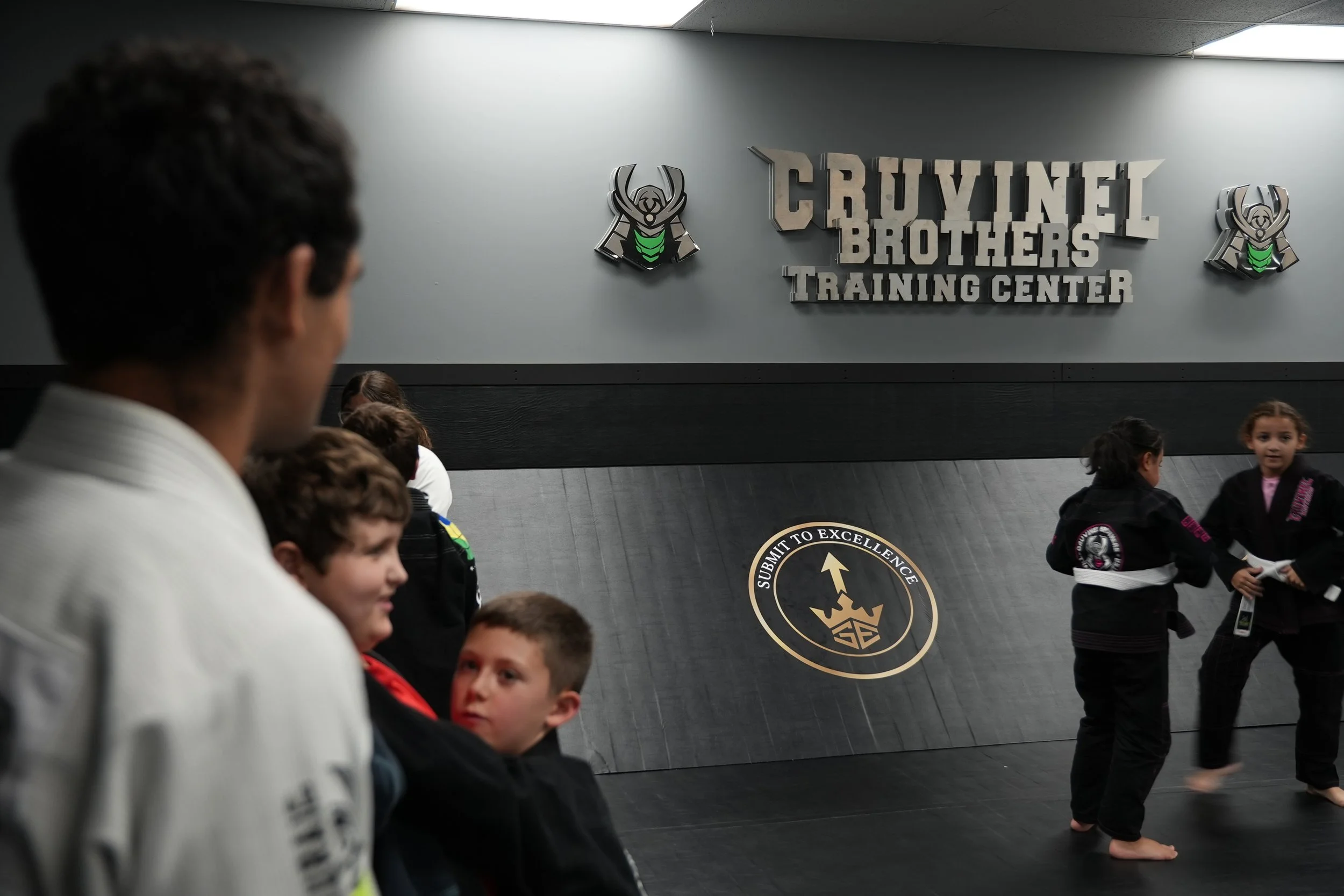 A group of children in martial arts uniforms standing in a training center with a sign that reads 'Crucial Brothers Training Center' and a logo of a black and green insect-like emblem.