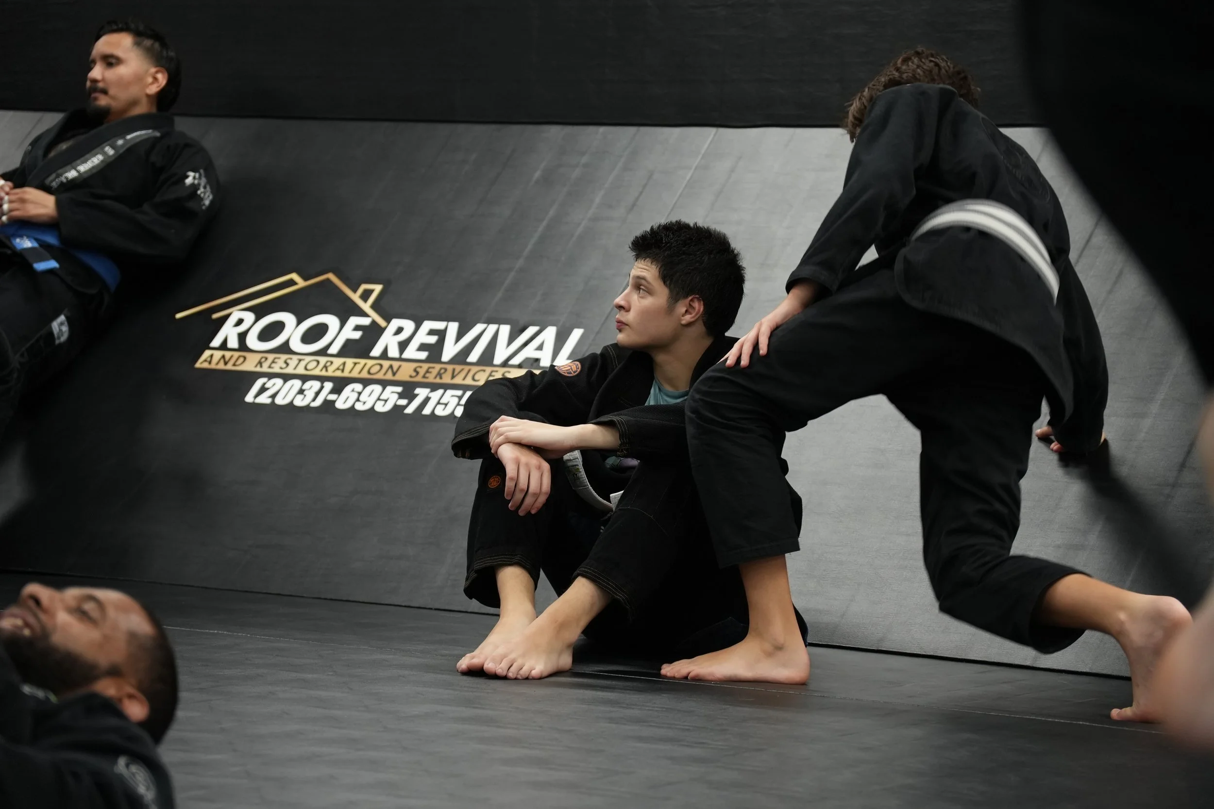 People practicing Brazilian Jiu-Jitsu on a black mat at a gym, with a sign that reads "Roof Revival and Restoration Services" on the wall.