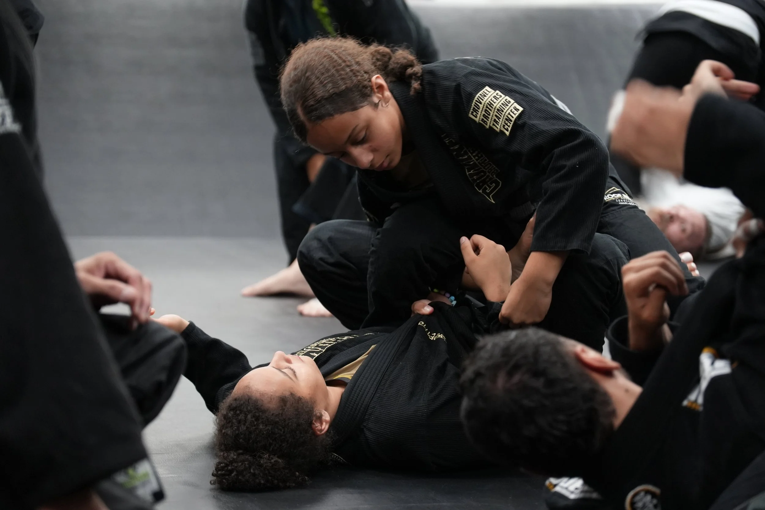 A group of people practicing Brazilian Jiu-Jitsu in a martial arts gym, with one person demonstrating a technique on another.