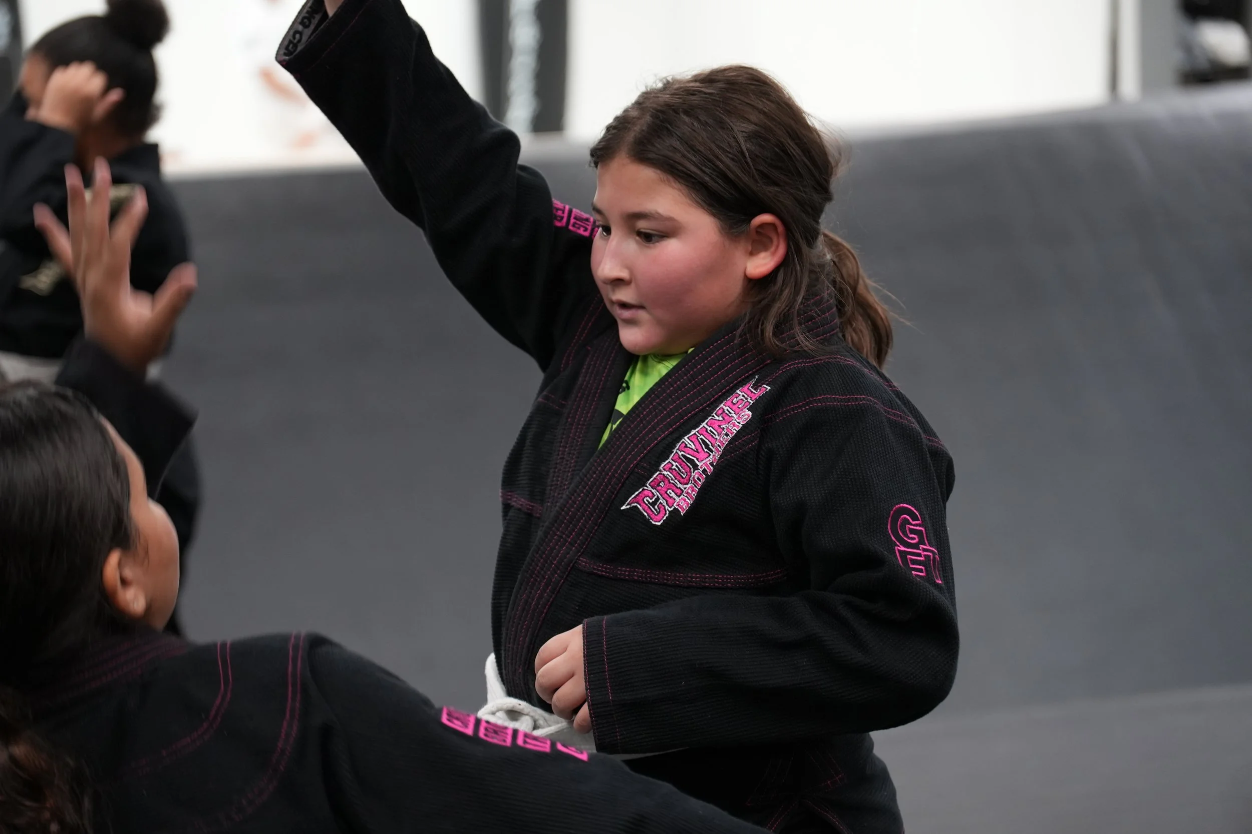 Children practicing Brazilian Jiu-Jitsu in a martial arts class, wearing black gi uniforms with pink and white logos, in an indoor gym or dojo setting.
