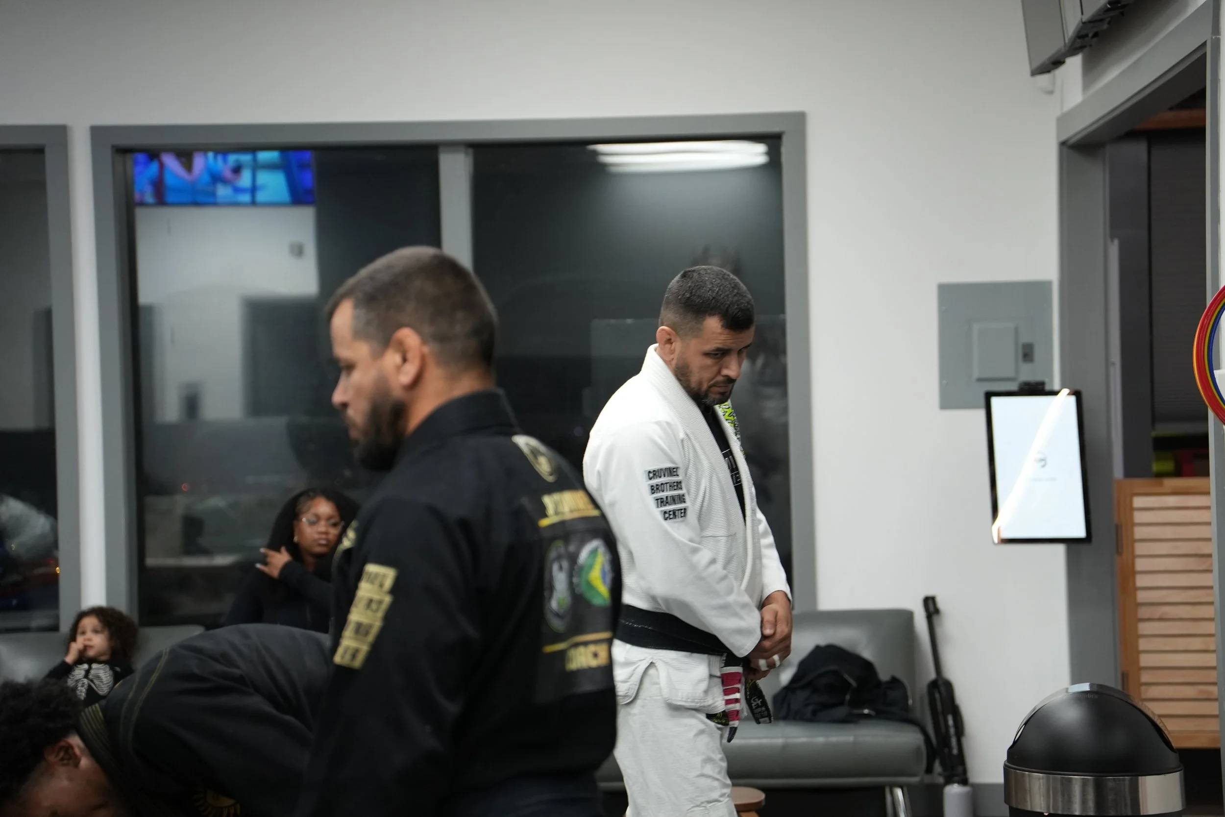 Two men standing in a room, one wearing a black uniform with patches and the other wearing a white martial arts gi, appearing to pray with heads bowed. In the background, a woman and a child are seated, and a woman is standing near the window. The ro