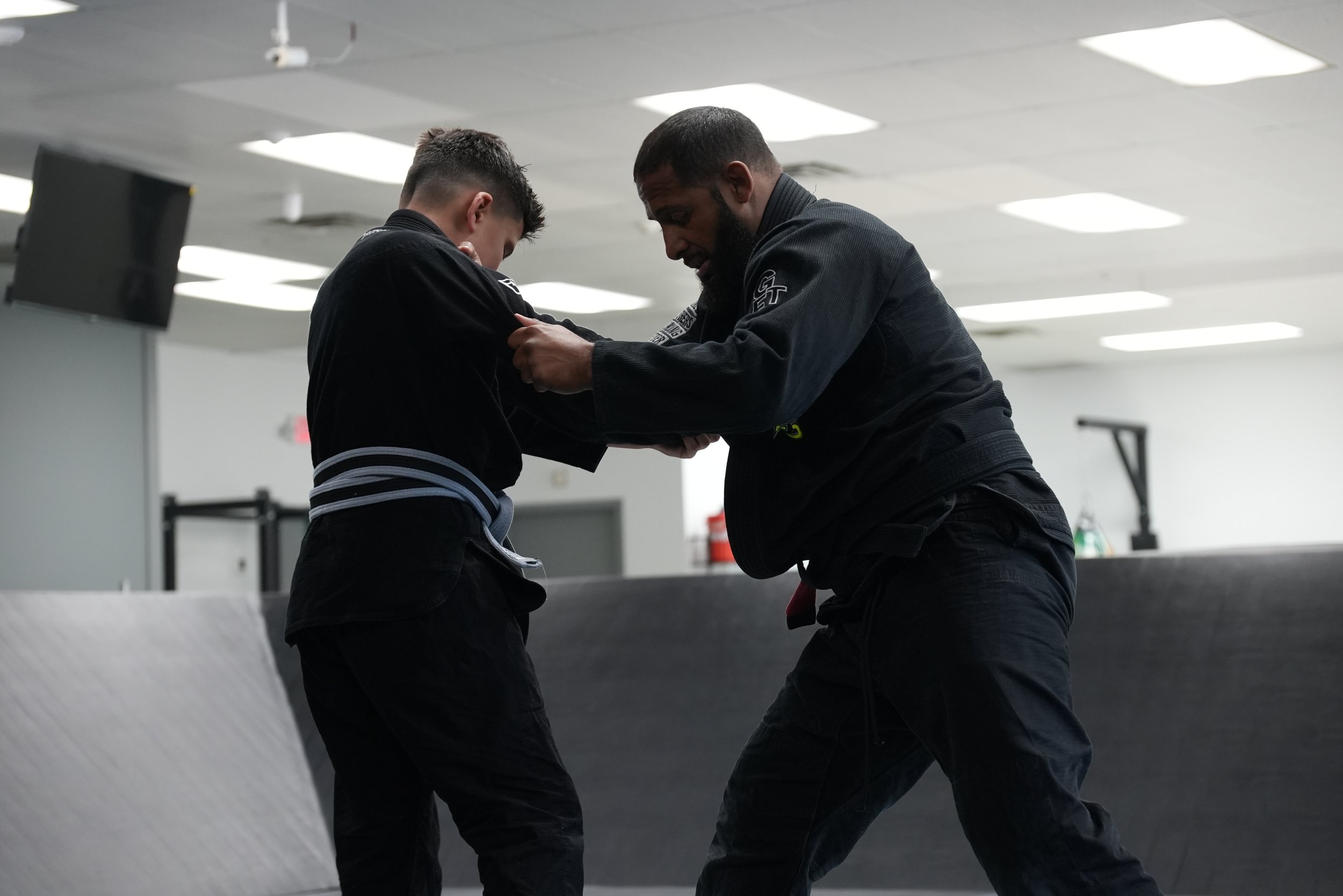 Two men dressed in black Brazilian Jiu-Jitsu gis practicing a technique in a training facility. One man appears to be helping or coaching the other.