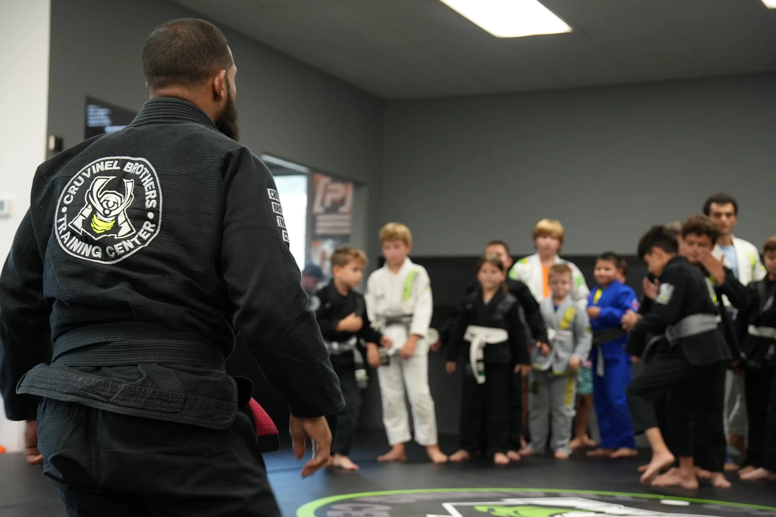 A martial arts instructor, wearing a black gi with a patch reading 'Cruvinel Brothers Training Center,' addresses a group of children in a martial arts class inside a dojo. The children are in gi uniforms, standing or sitting attentively.