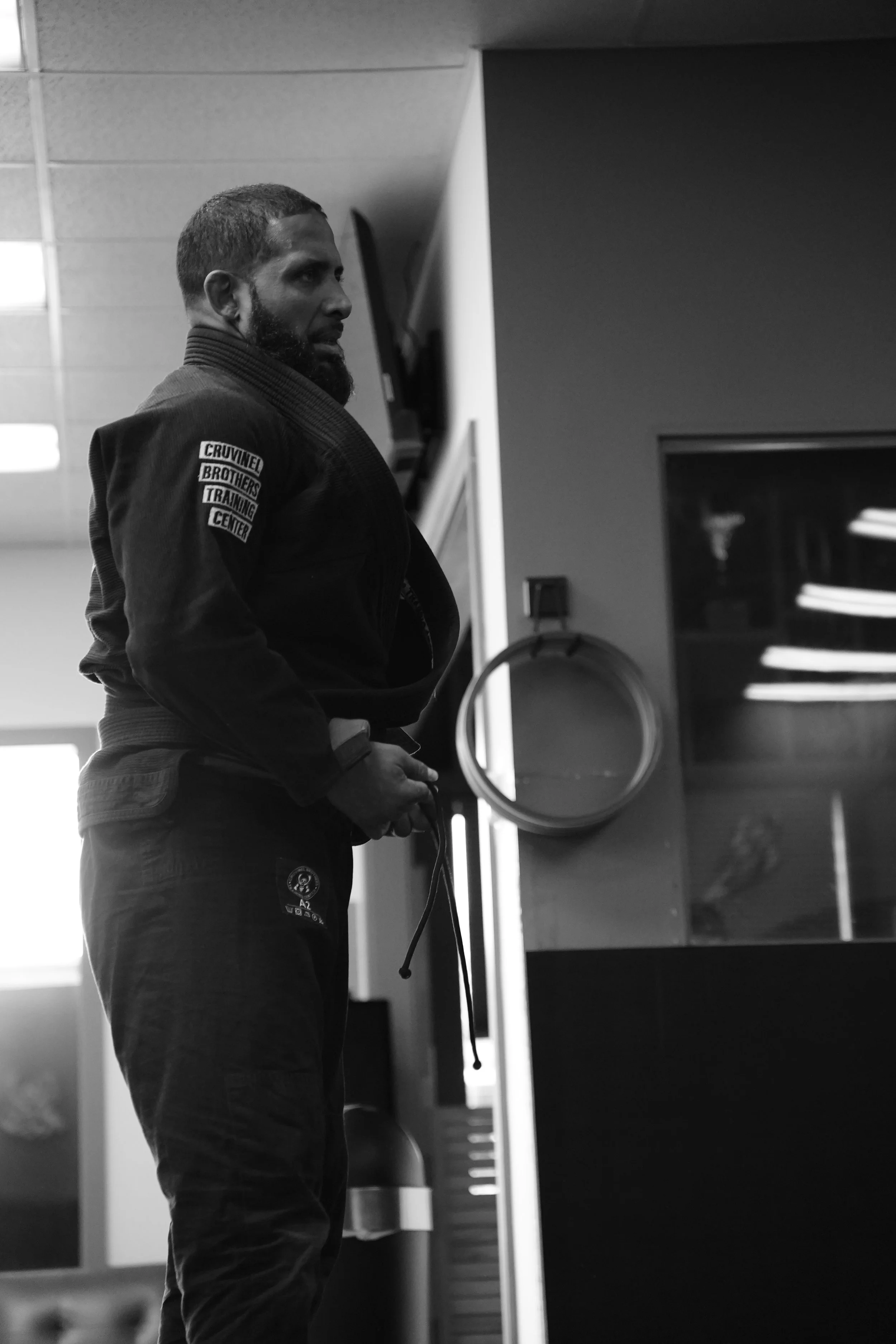 A man dressed in a Brazilian Jiu-Jitsu gi, standing indoors with his arms crossed, in a martial arts training facility.