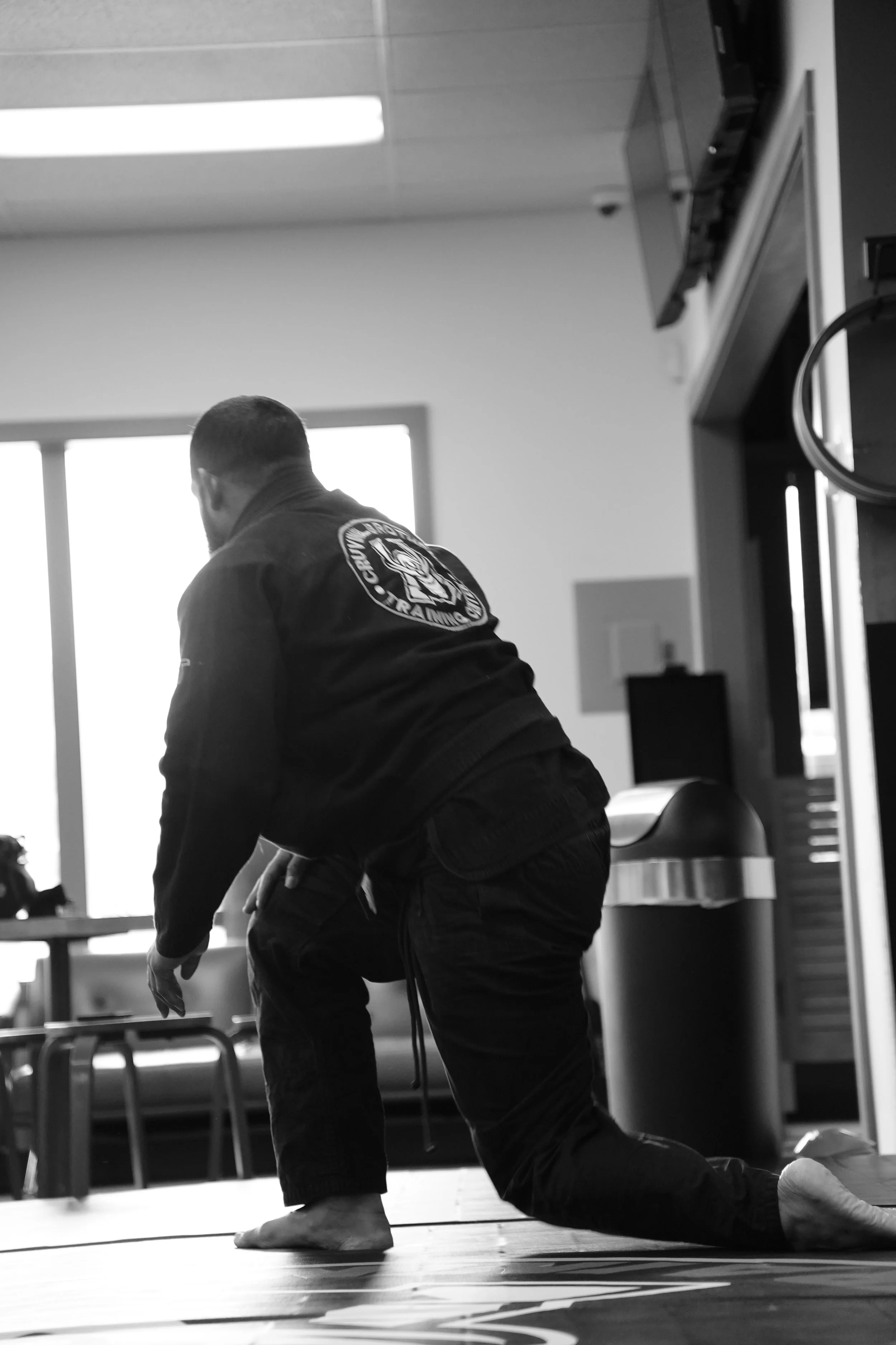 A person practicing martial arts or performing a martial arts move indoors, wearing a gi with a martial arts patch on the back, kneeling or in a bent over position on a mat.