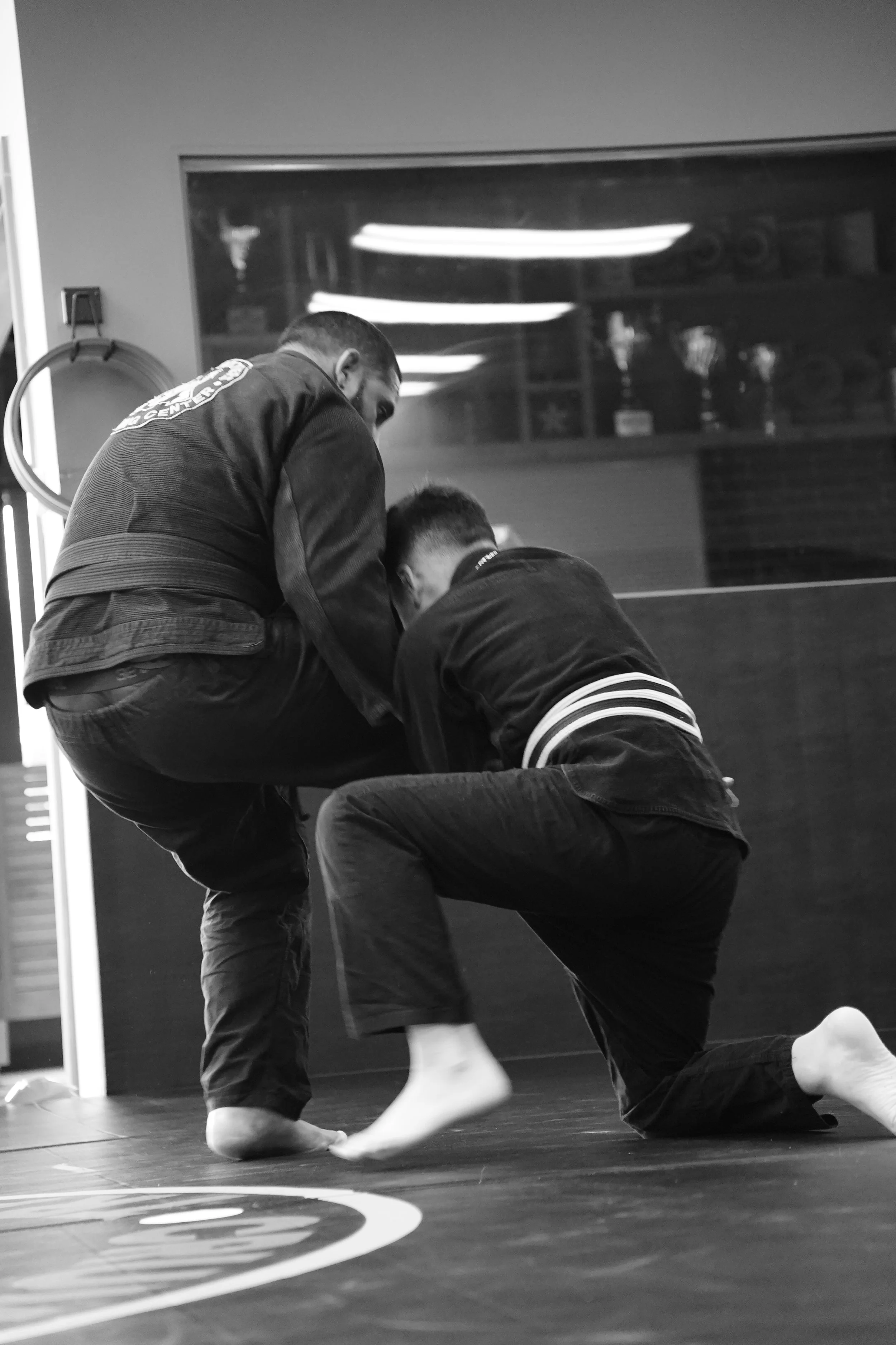 Two individuals practicing Brazilian Jiu-Jitsu on a mat, with one person kneeling and the other leaning forward, both dressed in gis.