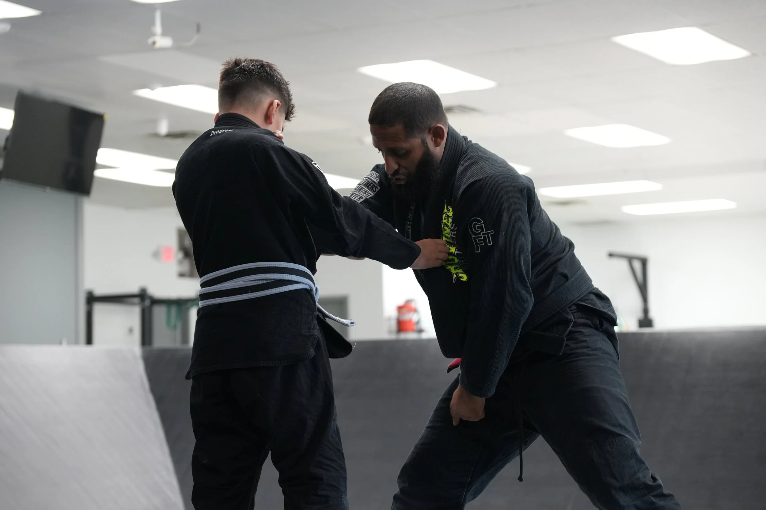 Two men training in a martial arts gym, one is wrapping the other's hand before a fight or training session, both wearing black martial arts uniforms.