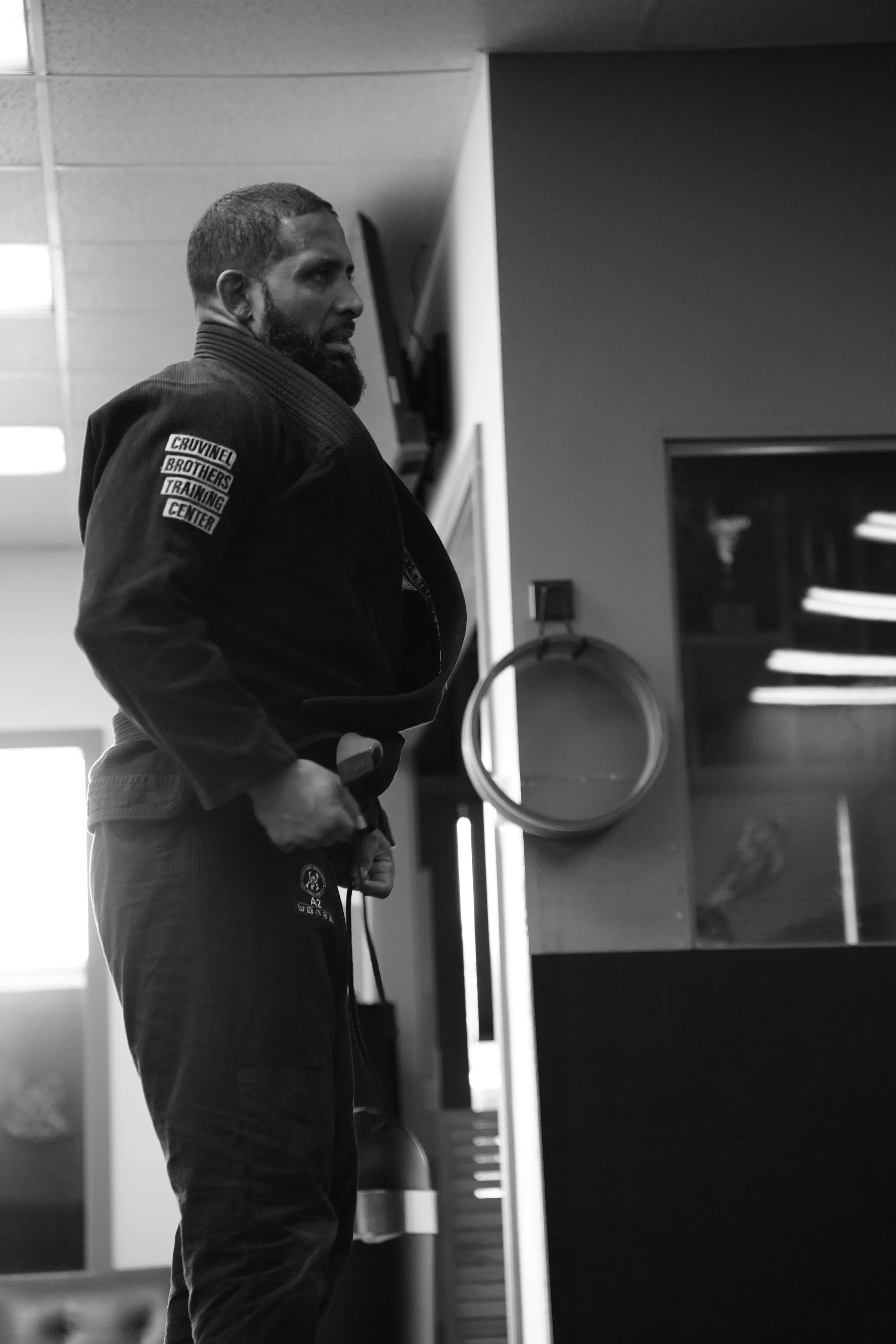 A man with a beard wearing a martial arts gi, standing indoors near a shelf with papers, holding a rope or cord, looking to the left.