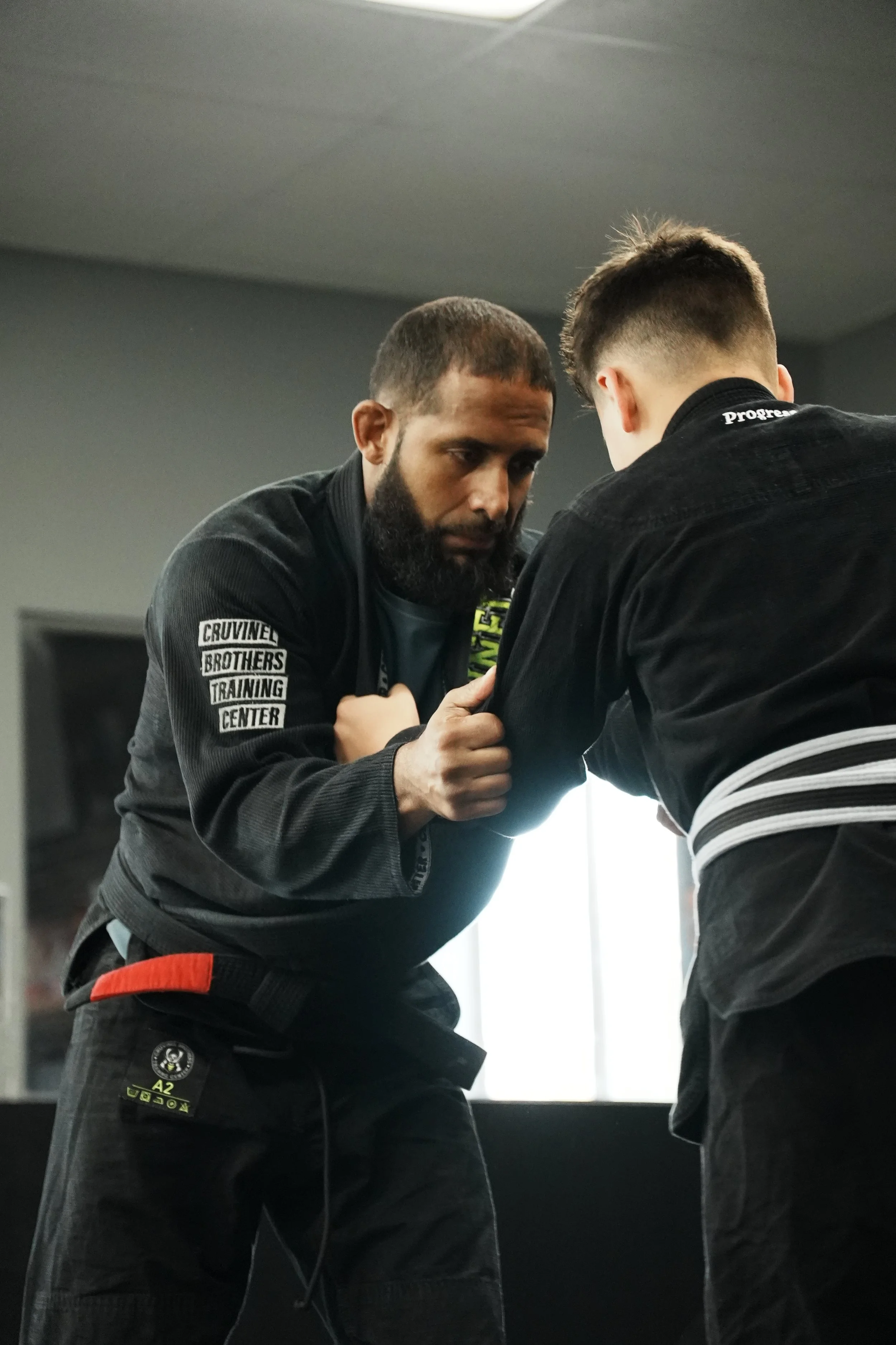 Two men are practicing Brazilian jiu-jitsu in a training facility. The man on the left has a beard and is wearing a black gi with patches on the sleeve that say "CRUVIANEL BROTHERS TRAINING CENTER" and other patches. The man on the right has short ha