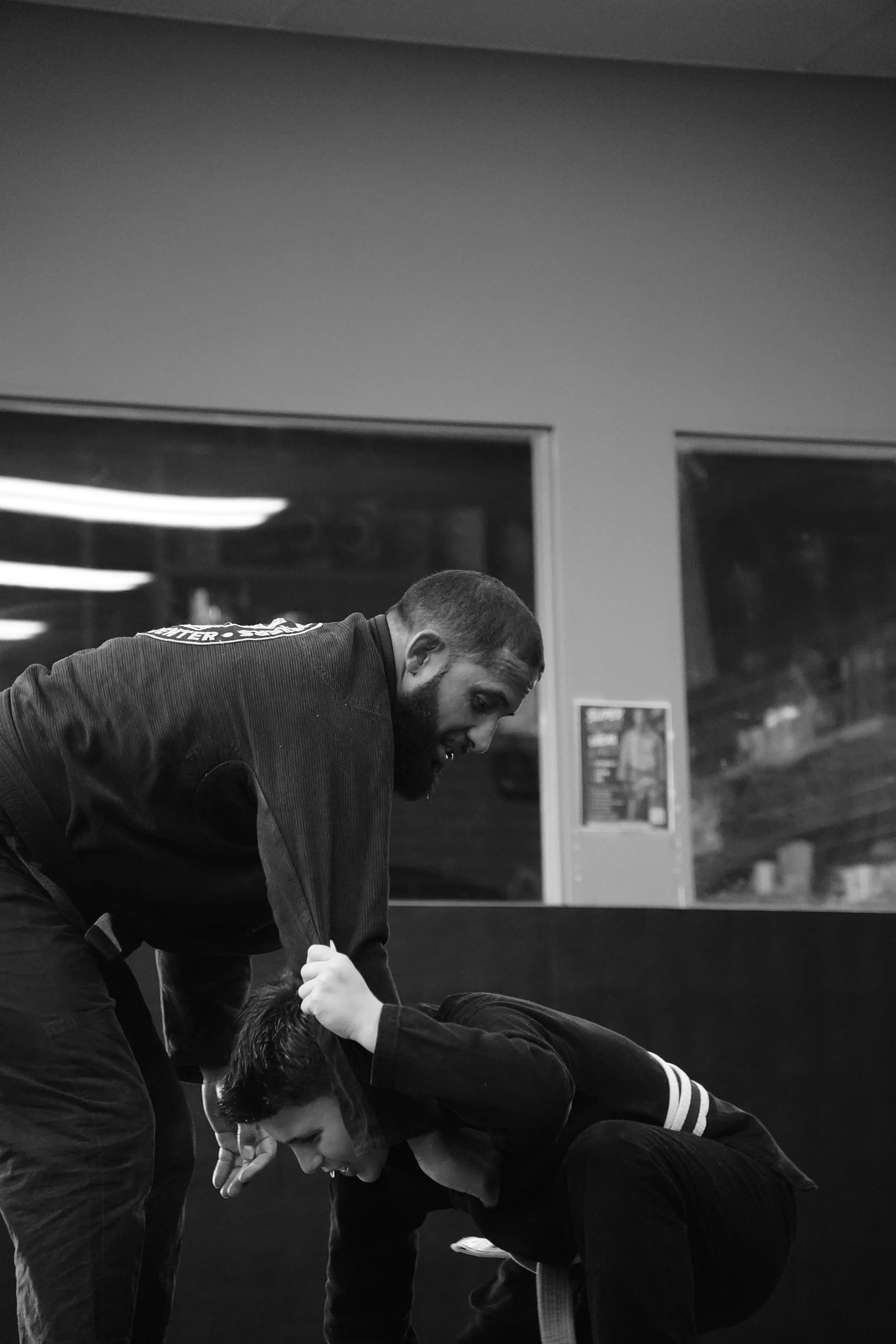 Two men practicing Brazilian Jiu-Jitsu in a gym, both are wearing Gis. One man is standing and the other is on the ground, with the standing man gripping the other's collar.