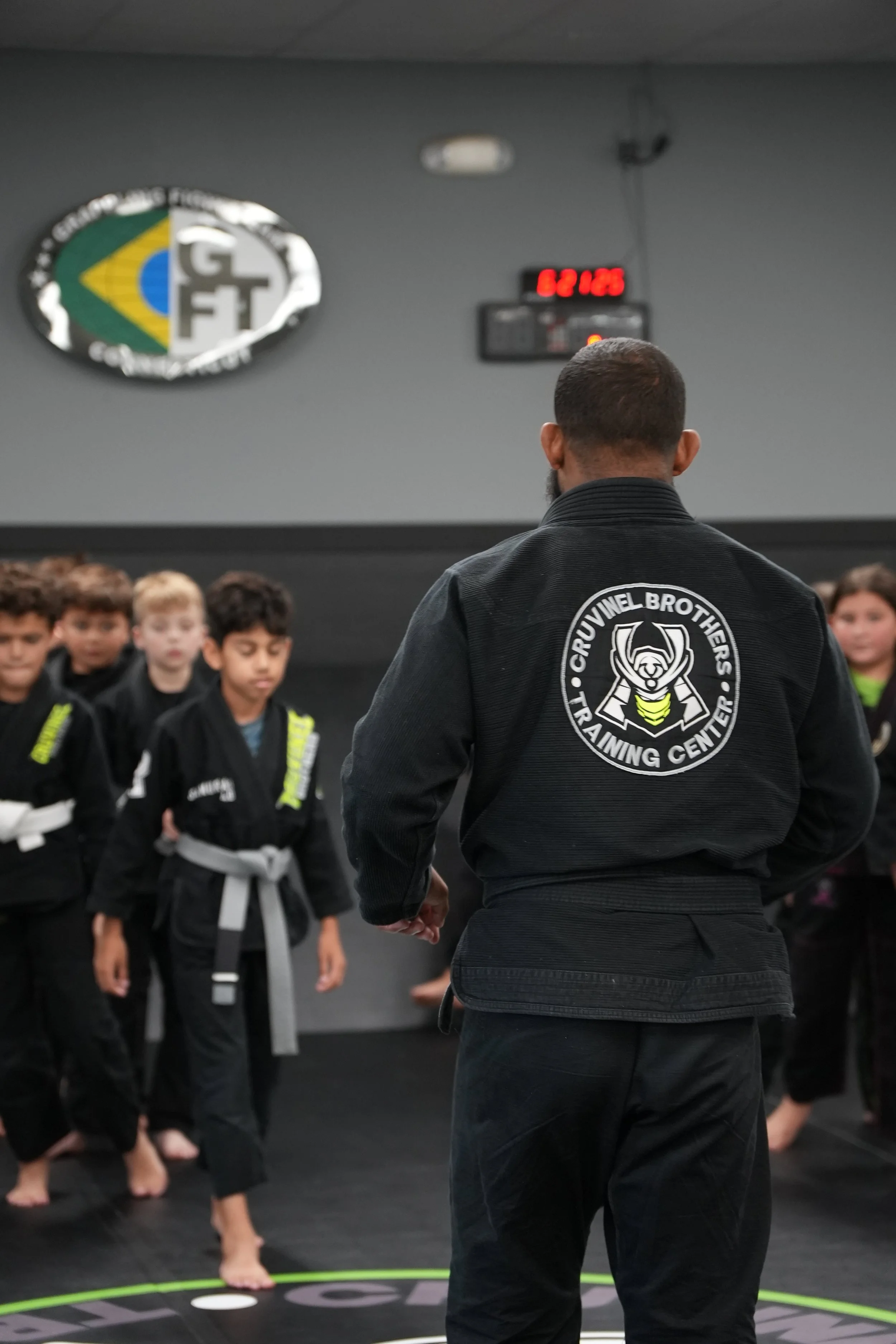 Martial arts instructor in a black uniform with 'Crivinel Brothers Training Center' logo teaches a group of young students in black uniforms at a martial arts training facility.