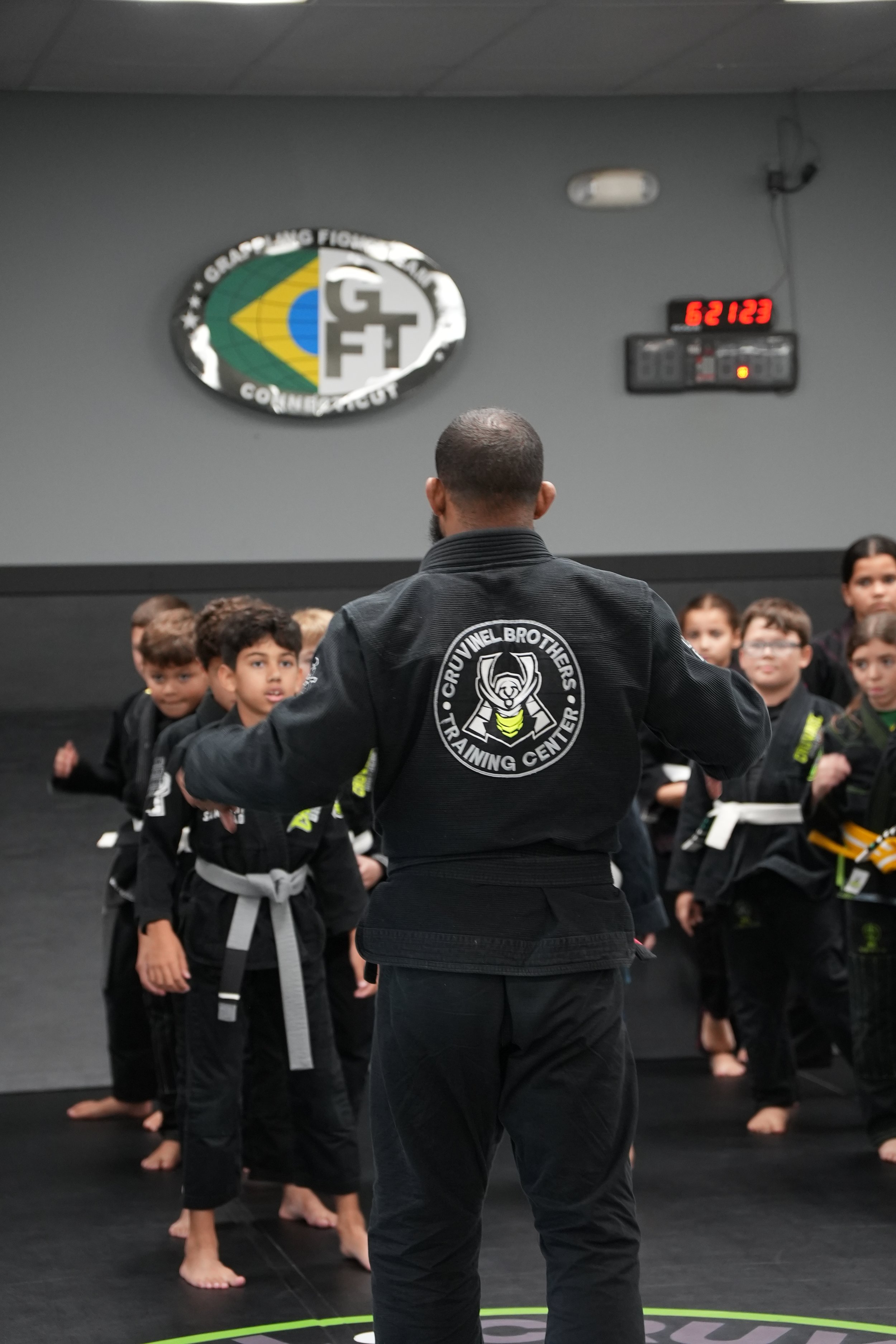 Black belt instructor with 'Cruzuel Brothers Training Center' logo on their gi, standing in front of a group of young students in martial arts training uniforms, inside a dojo with Brazilian flag logo and a digital clock on the wall.