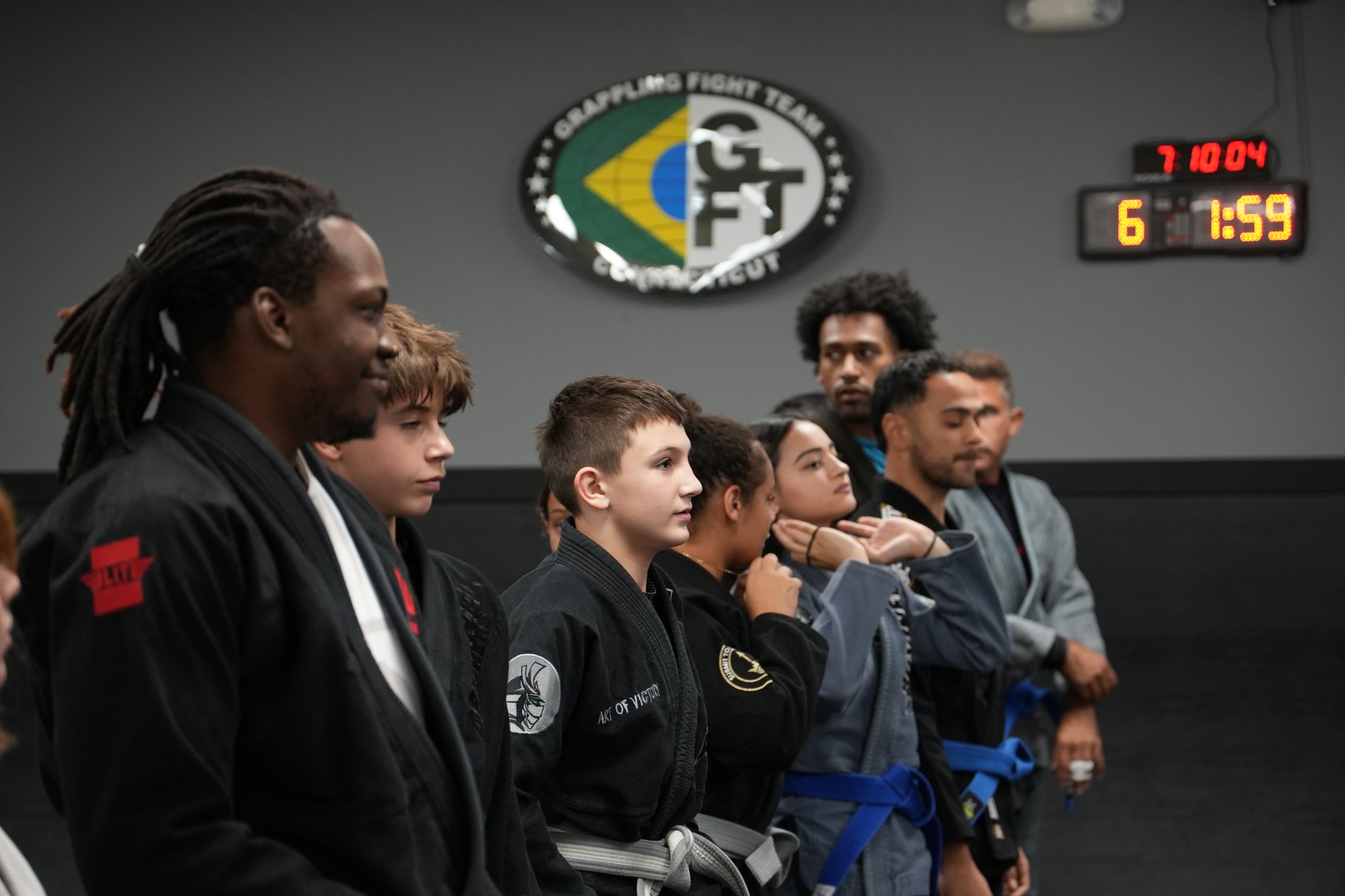 Group of martial artists standing in a line with their eyes closed, wearing martial arts uniforms, inside a dojo with a logo on the wall and a digital clock showing 7:10 am.