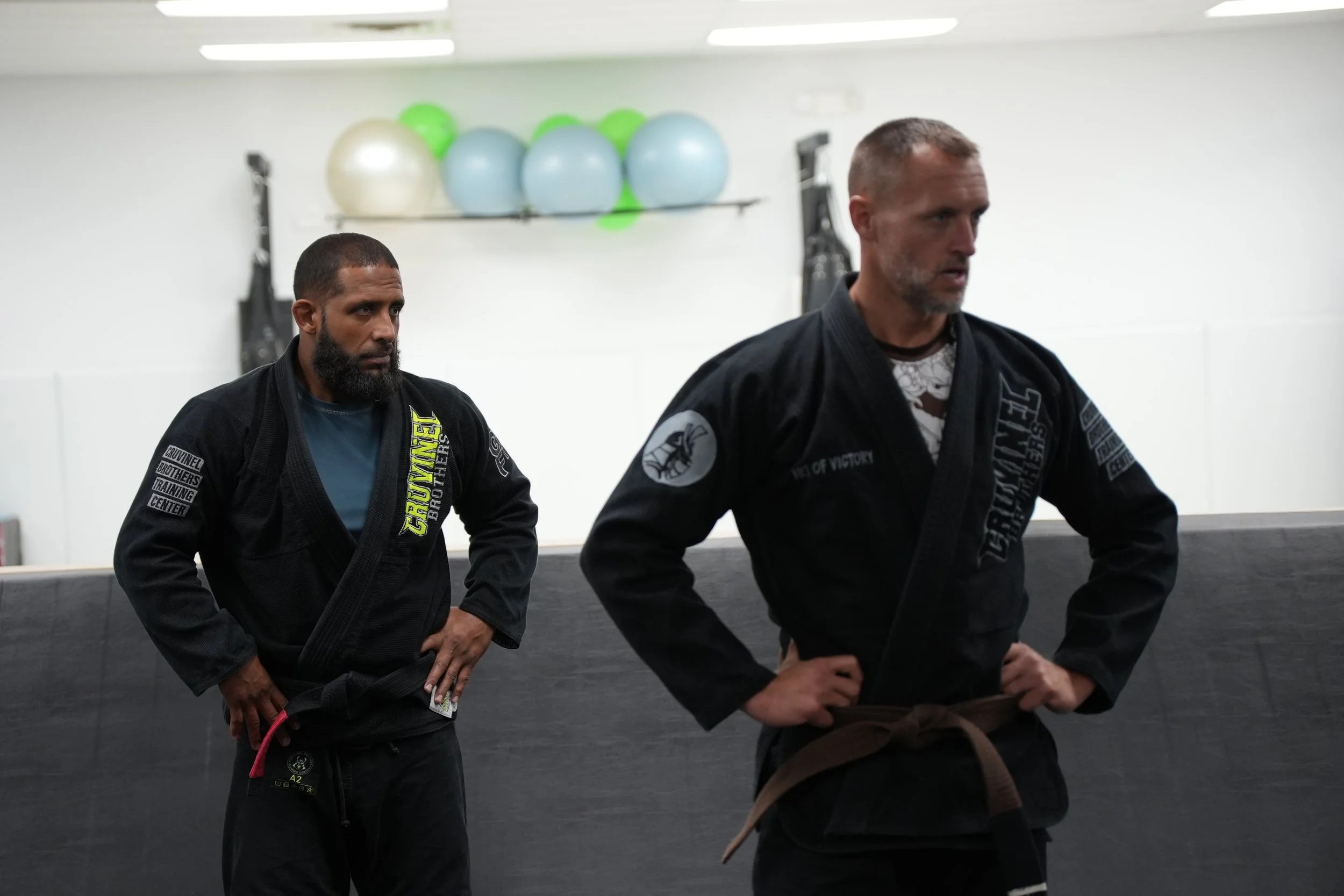 Two men in black jiu-jitsu gi uniforms standing in a martial arts training facility, with exercise balls on a shelf in the background.