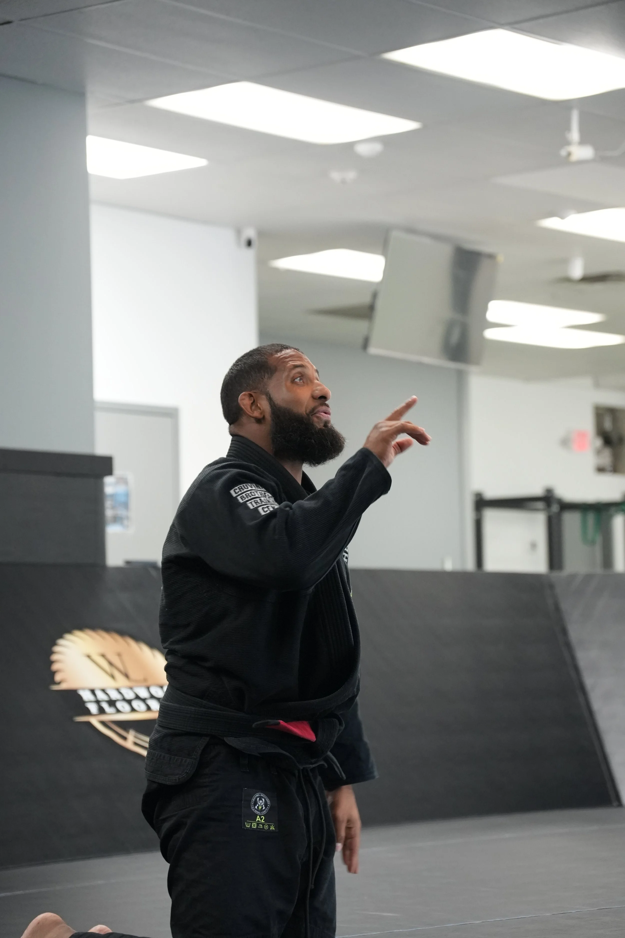 A man in a black Brazilian Jiu-Jitsu gi with patches stands in a gym, pointing upward with one hand, with a partially visible instructor or teammate nearby. The background includes a black wall with a logo, mirrors, and ceiling lights.