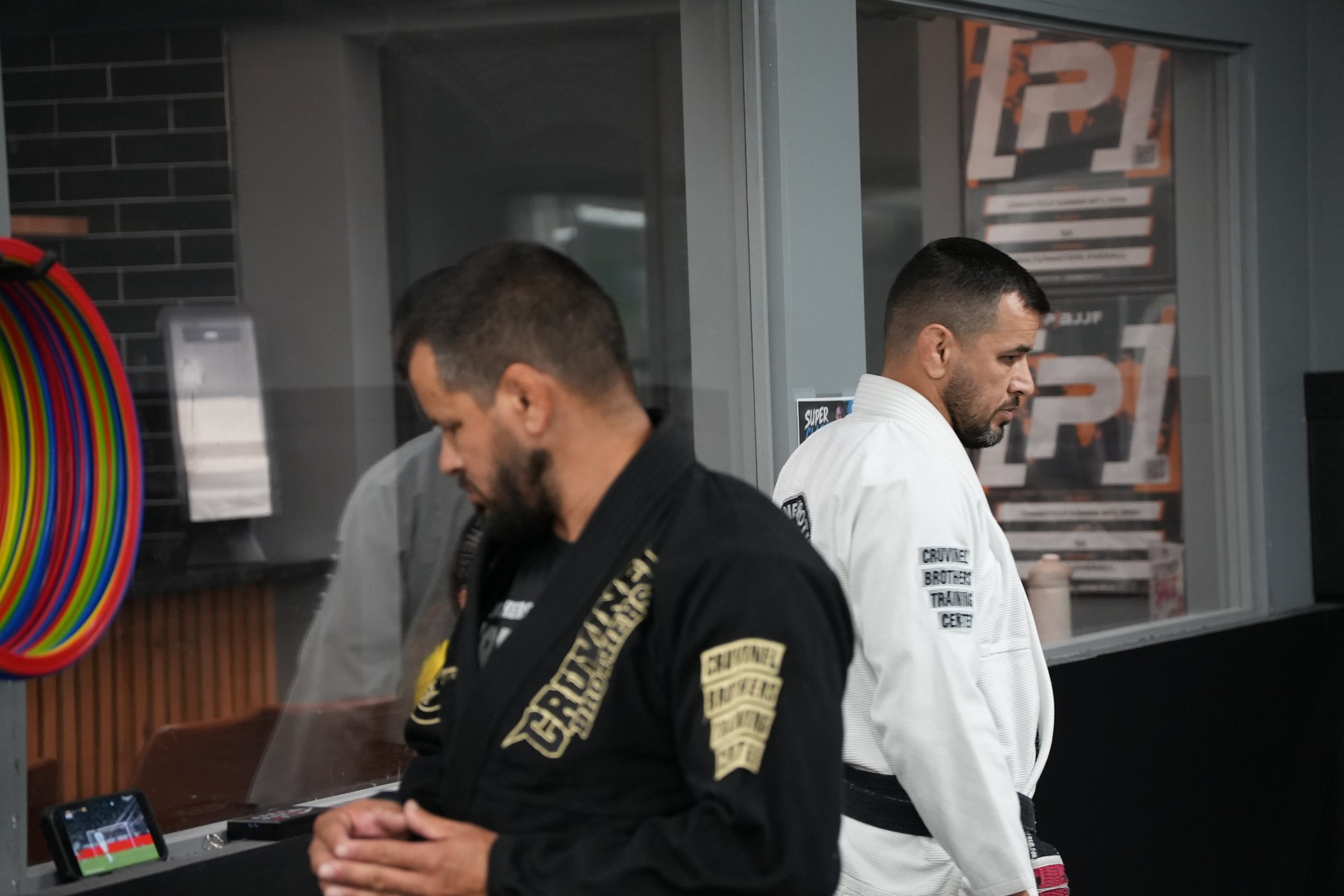 Two men in martial arts uniforms standing inside an indoor dojo training center, one wearing a black gi and the other in a white gi, with a colorful hula hoop and training notices in the background.