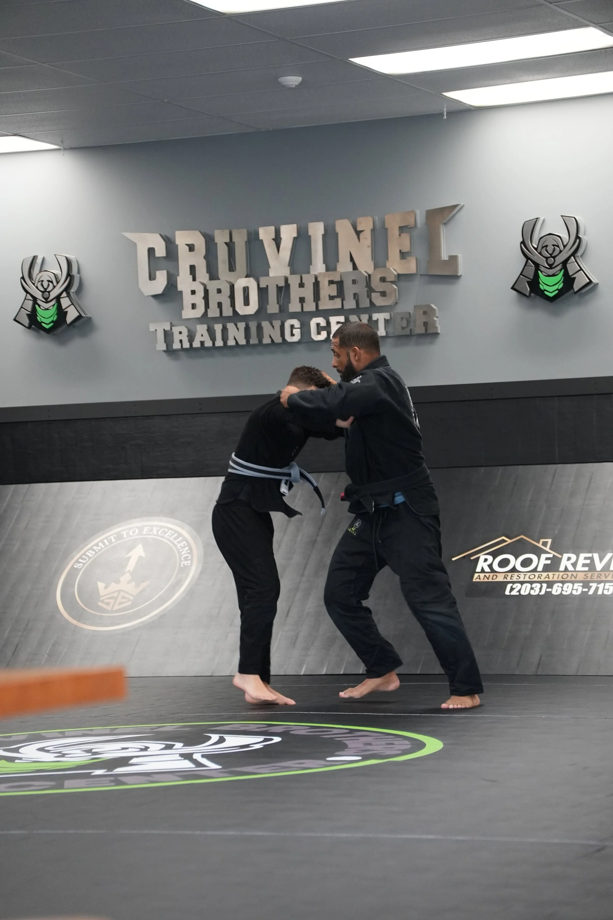 Two people practicing Brazilian Jiu-Jitsu on a black mats with a logo, inside a gym with gray walls. The person on the left is wearing a gi with a black belt, and the person on the right is in a black gi. They are engaged in a grappling match.