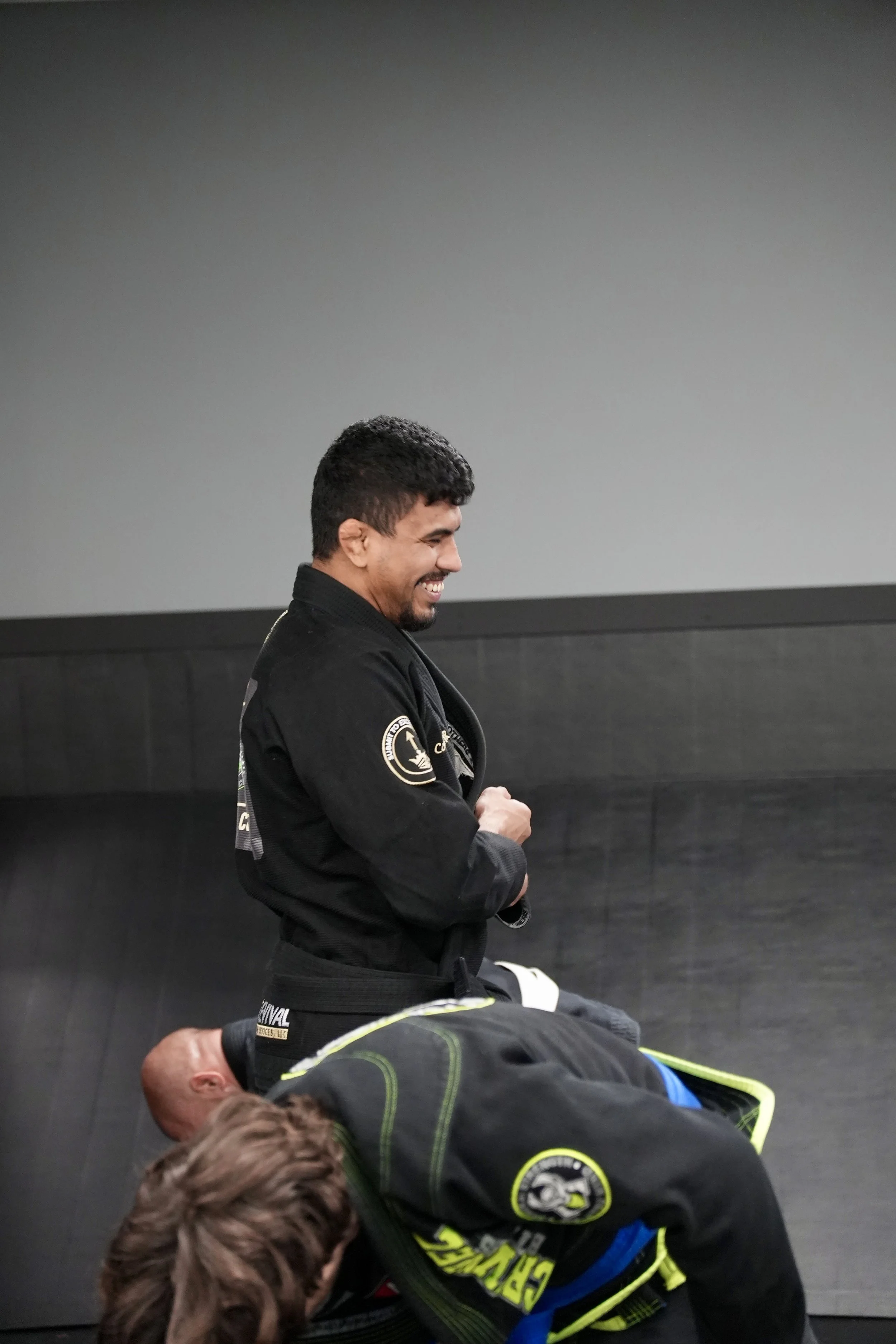 A group of martial artists practicing in a dojo with a gray wall, with some standing and others in a kneeling position.