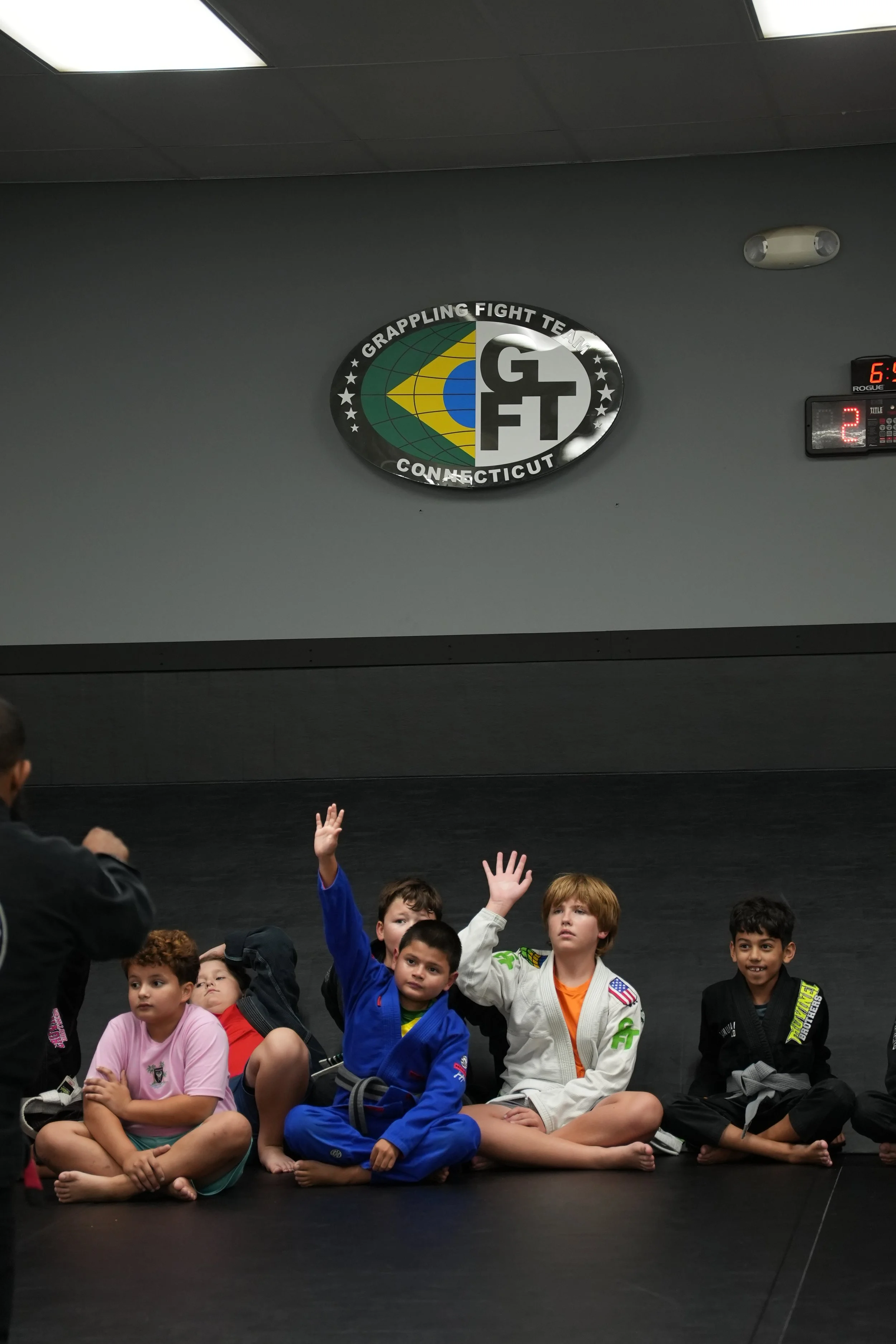 Group of children sitting on the floor in a martial arts gym, some raising their hands, with a gray wall and martial arts logo behind them.