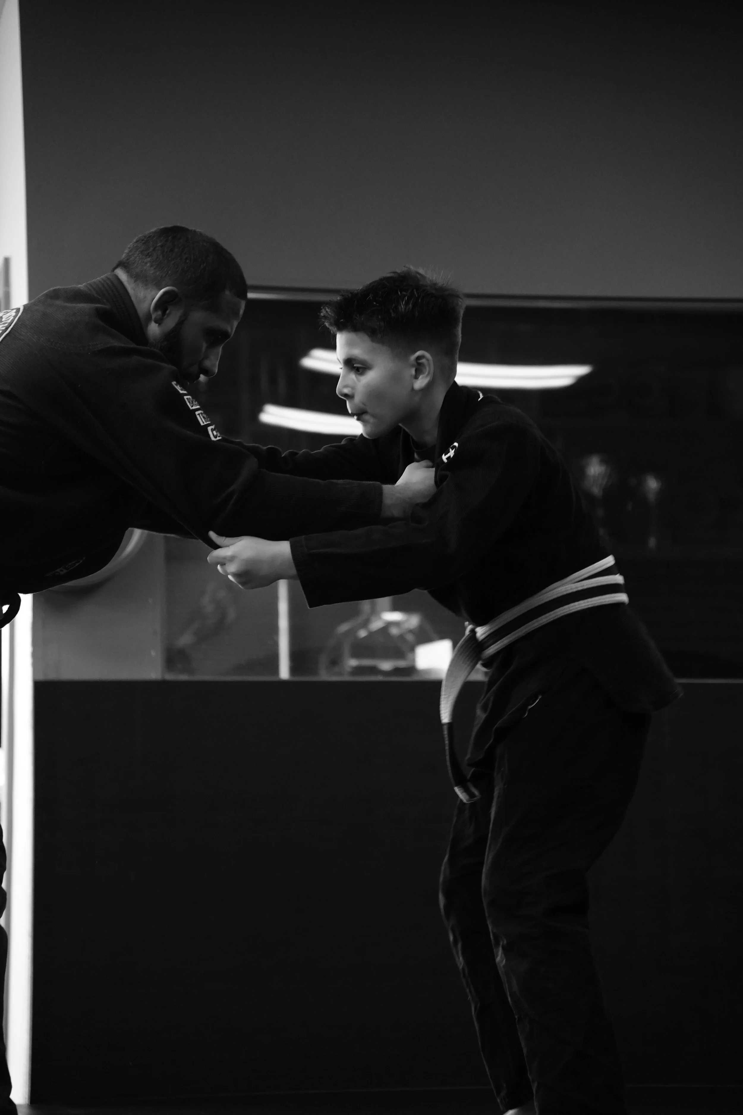 A black and white photo of a martial arts instructor teaching a young student how to perform a technique, both wearing traditional martial arts uniforms, in a dojo or training facility.