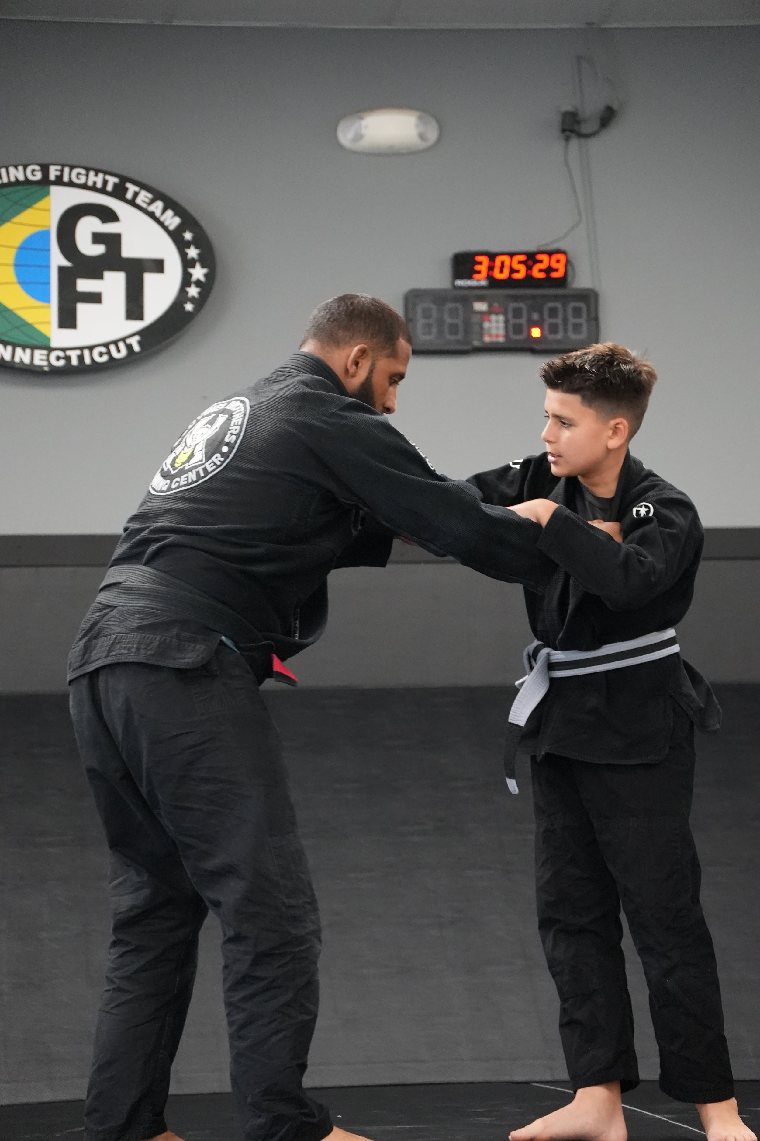 Two martial artists training in a gym, wearing gis, with a black wall and a digital clock showing 3:05:29 in the background.