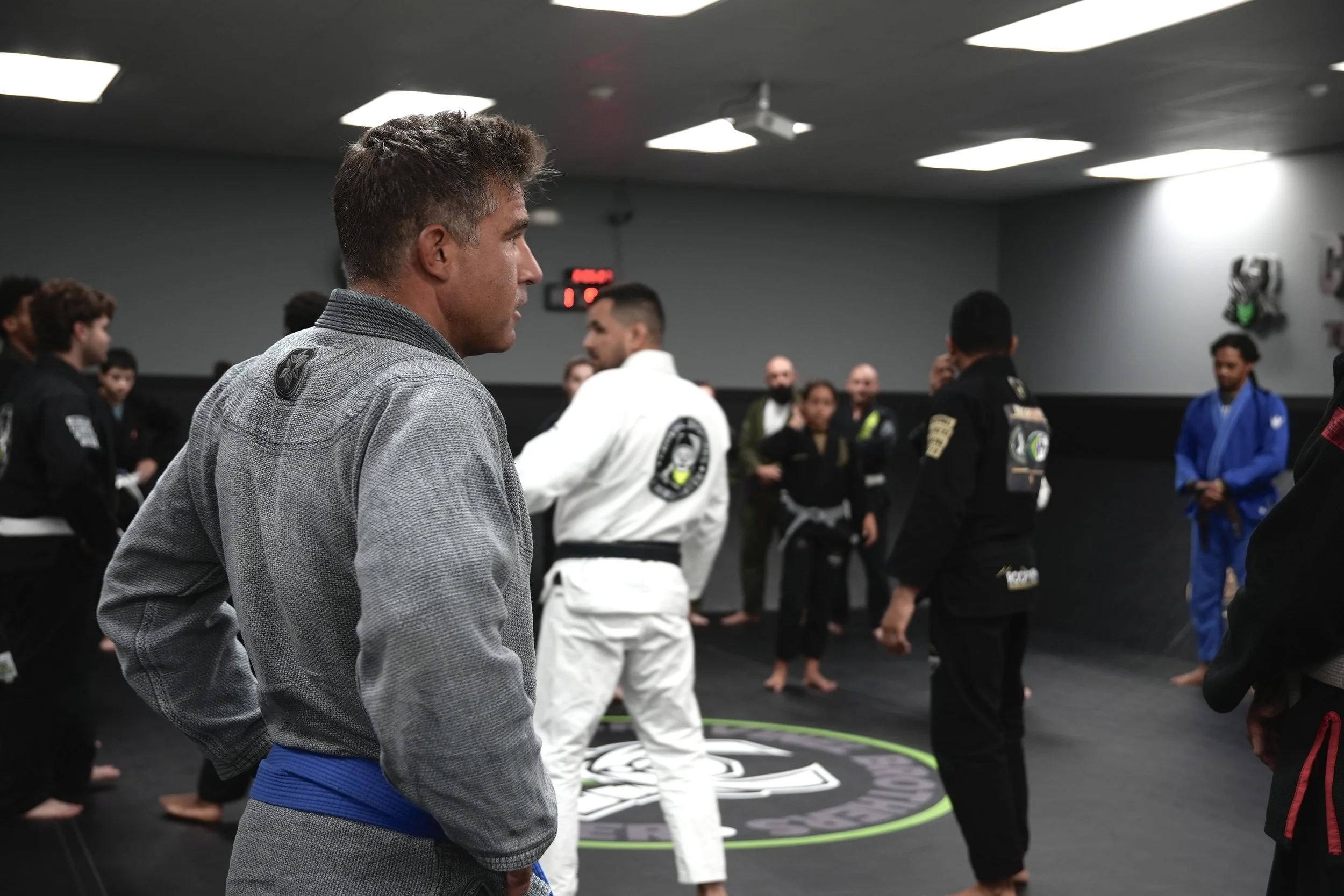 A group of martial artists in gis and belts gathered in a dojo or martial arts training center, standing in a circle or semi-circle, with a man in a gray gi with a blue belt in the foreground, possibly listening to instructions or observing.