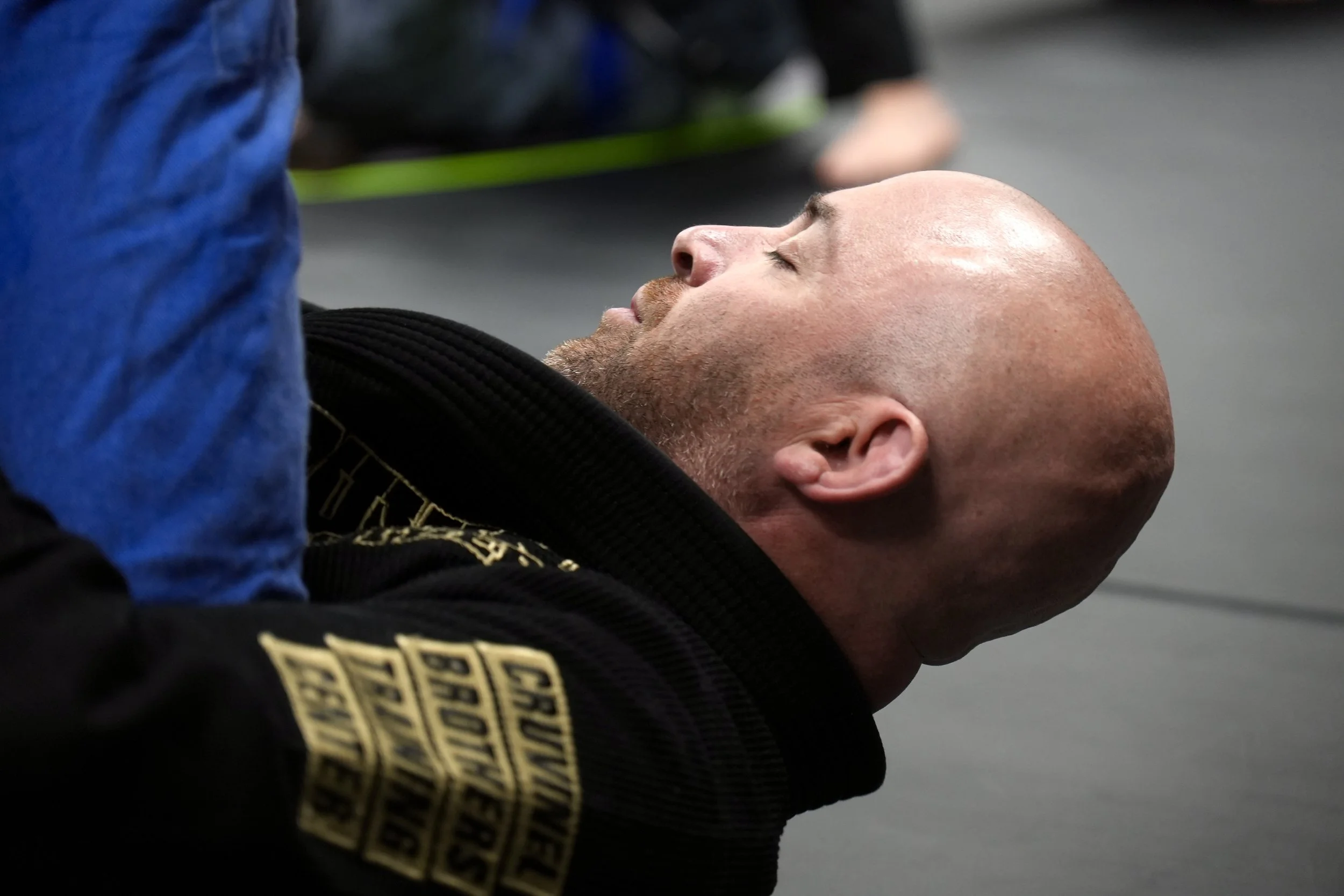 A man lying on his back on a mat with eyes closed, possibly resting or in a meditative state. He is wearing a black gi with a gold belt, indicating martial arts practice. Part of another person's arm and blue clothing are visible in the foreground.