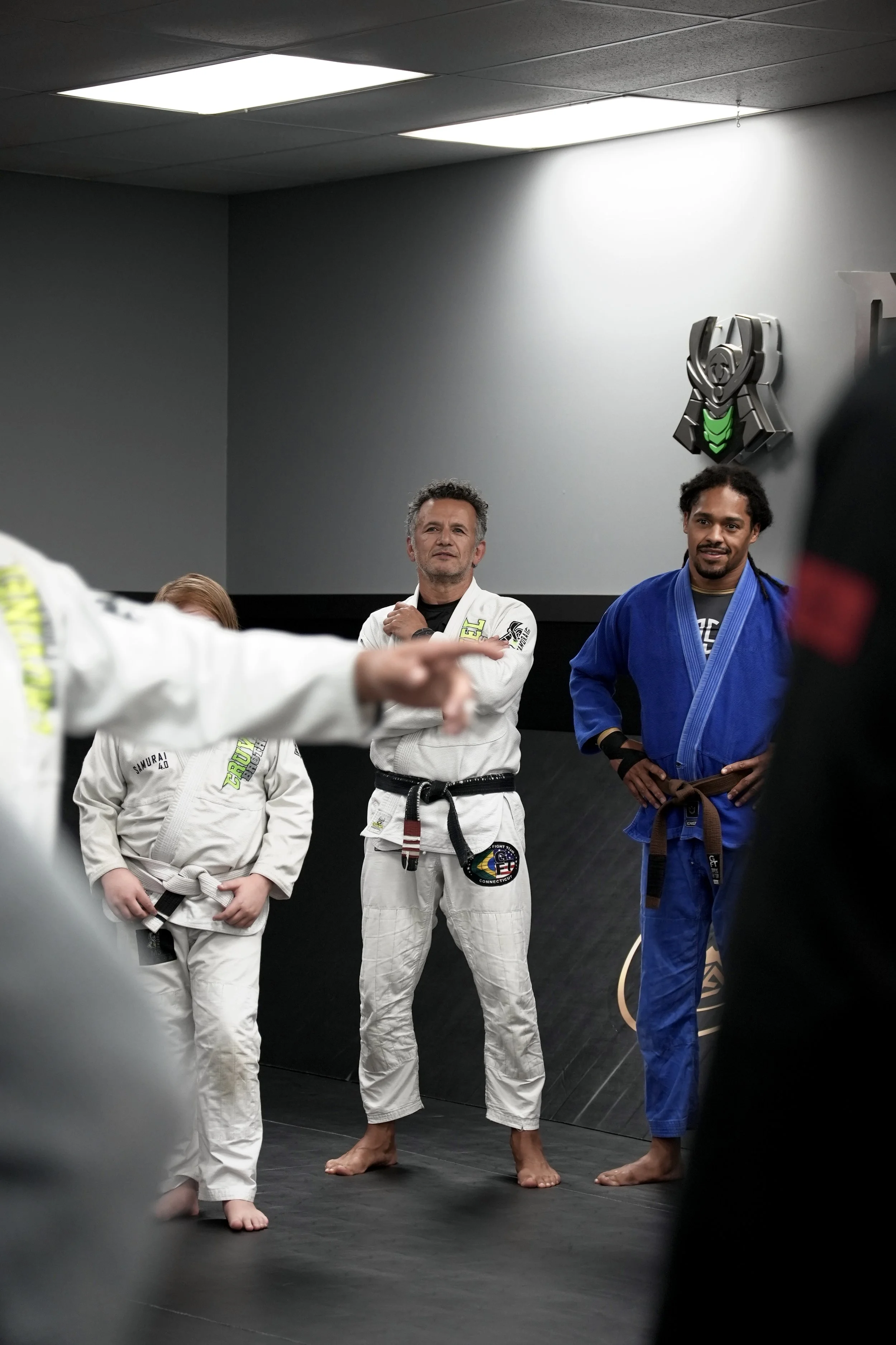 Brazilian Jiu-Jitsu practitioners in uniform, standing in a dojo with a gray wall and a martial arts emblem above them. Two men in white gis and a woman in a white gi with a black belt are visible, along with a man in a blue gi and brown belt, all ba