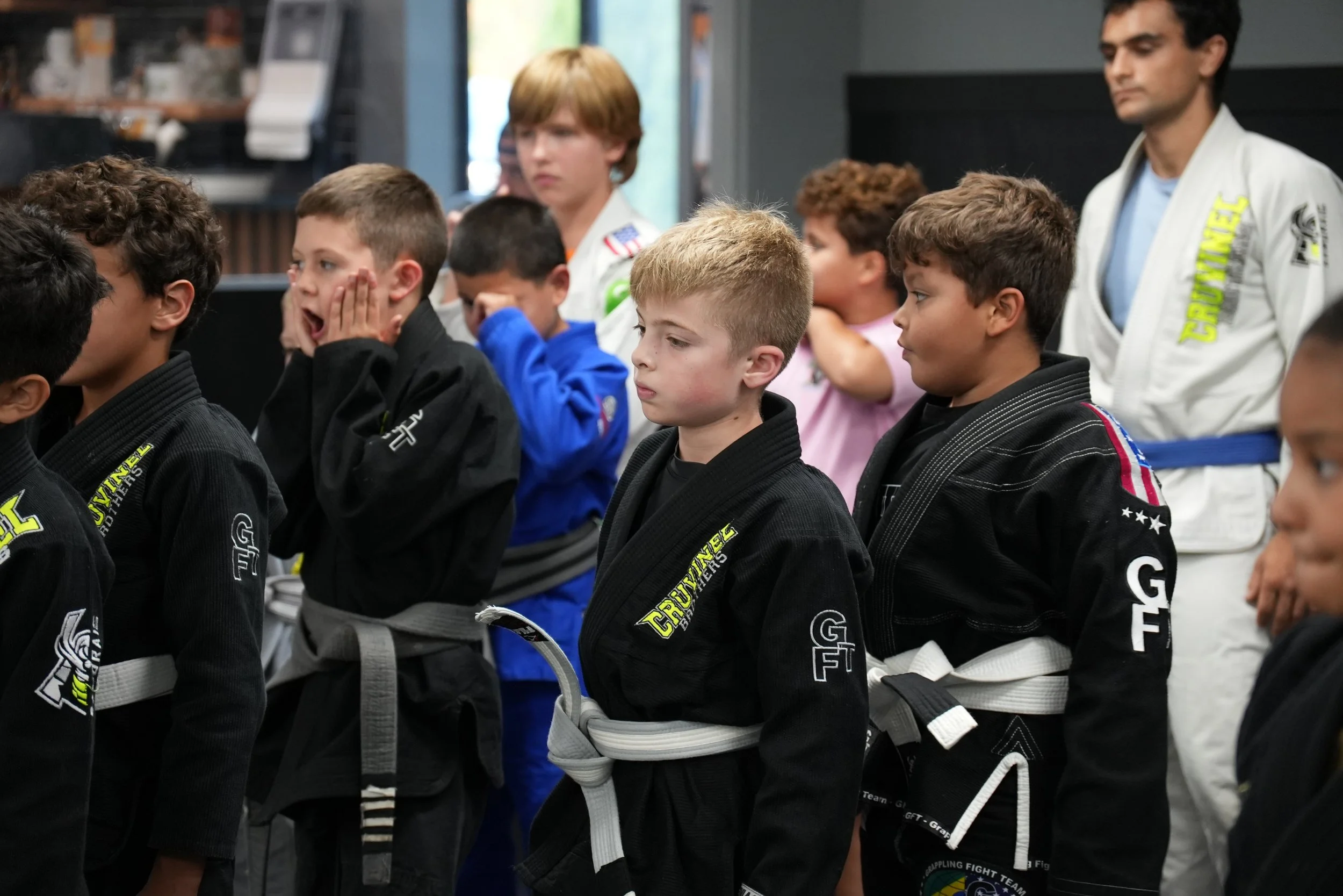 Group of children dressed in Brazilian Jiu-Jitsu gis standing in a training room, some adjusting their gis or listening.