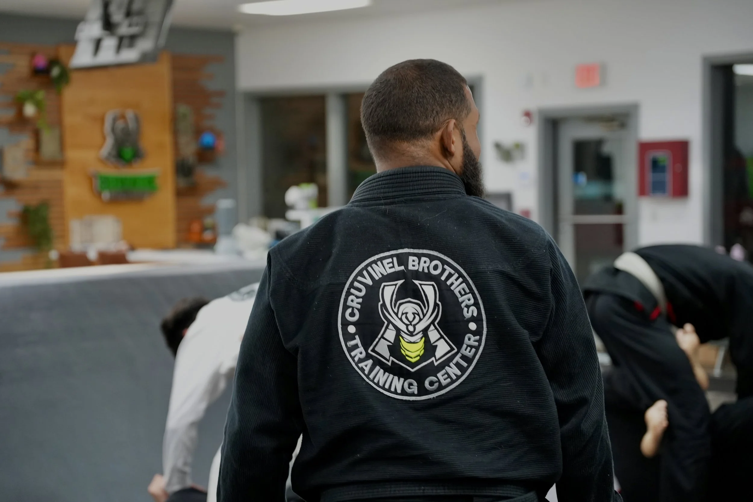 Martial arts class with students practicing moves, instructor in black gi with a 'Kruvinel Brothers Training Center' logo on back, in a room with gray walls and wooden decor.