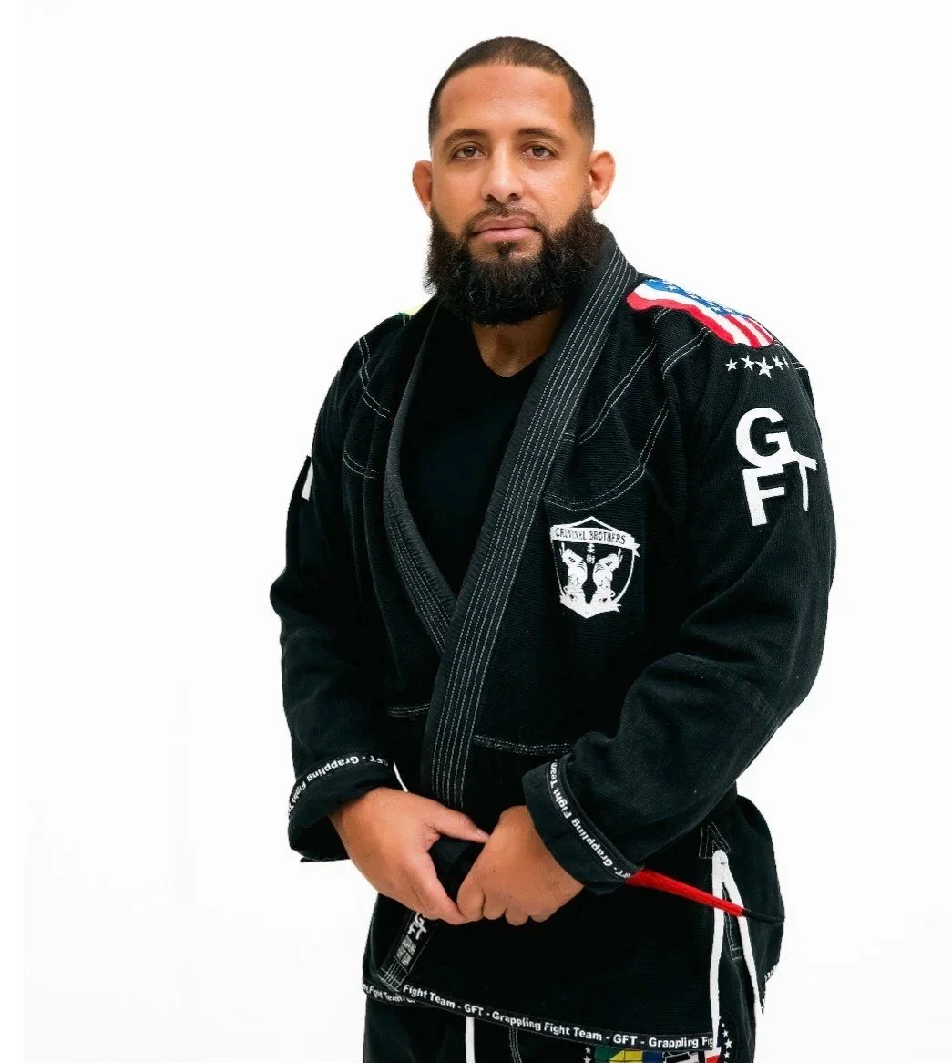 A man in a black Brazilian Jiu-Jitsu gi with patches and embroidery, standing against a white background.