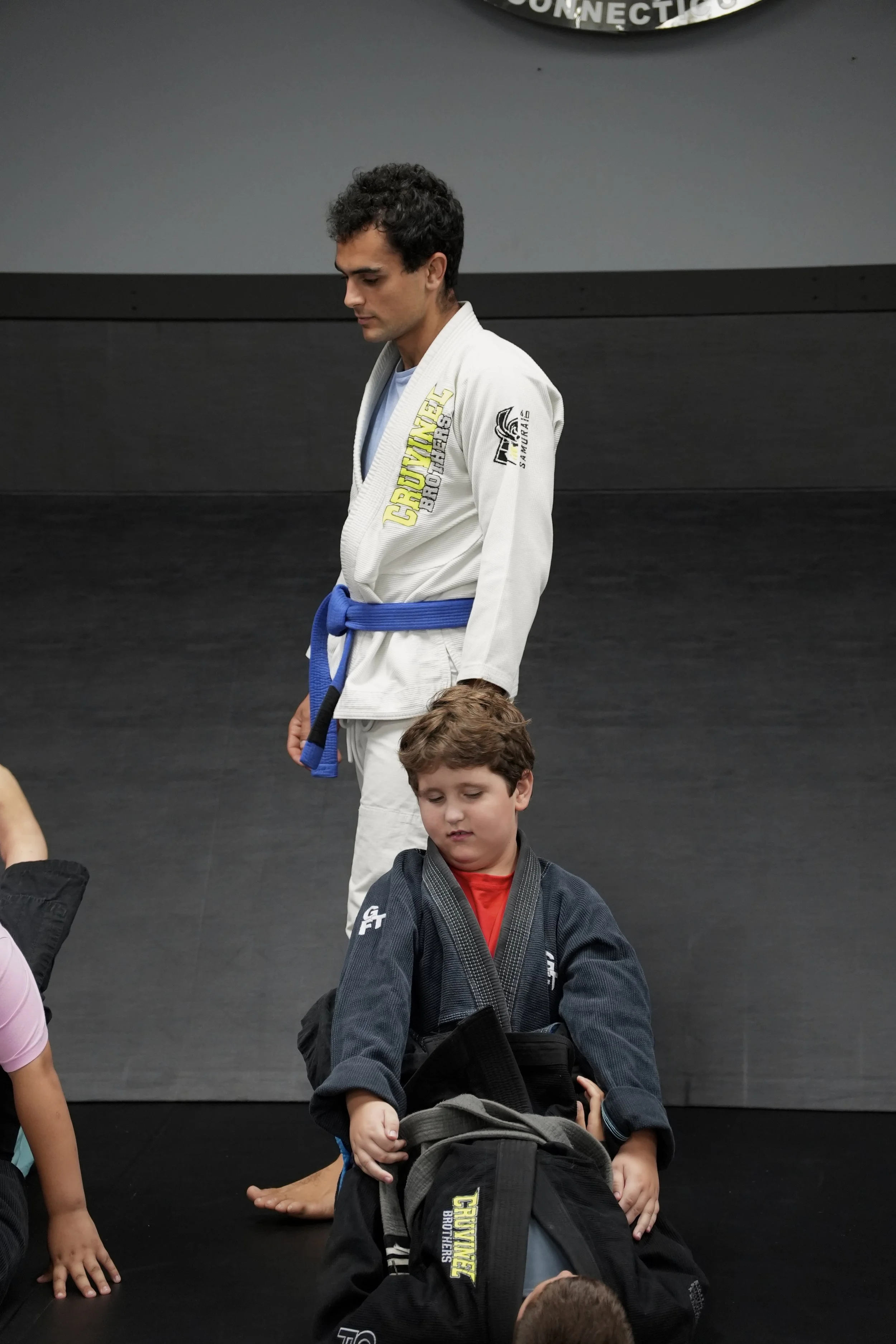 A man in a white Brazilian Jiu-Jitsu gi with a blue belt stands on a black mat in a martial arts studio, looking down. Several children wearing gis and black belts are seated or kneeling on the mat around him.