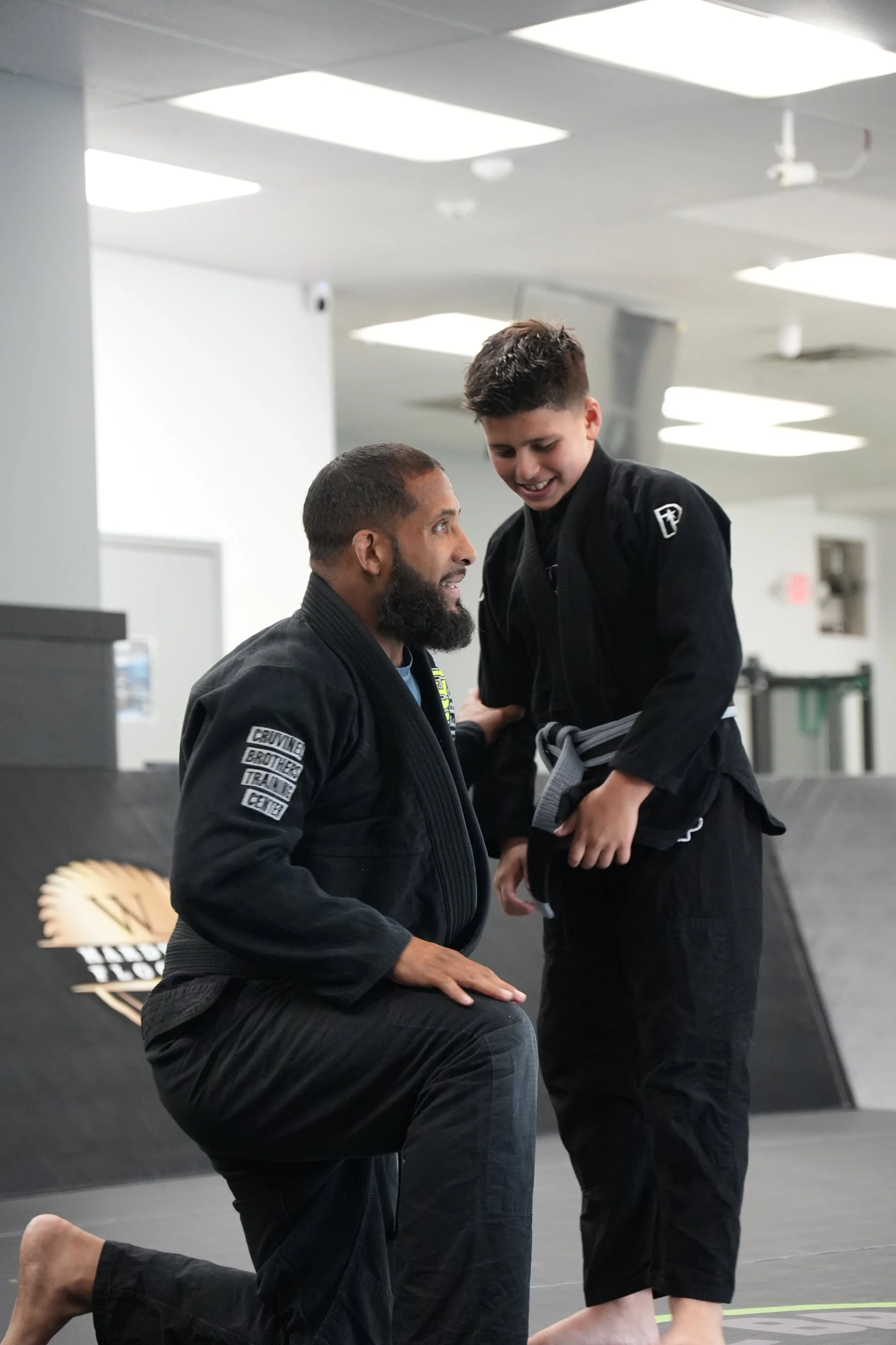 An instructor kneeling on one knee in a martial arts gym, speaking to a young student who is smiling and wearing a black gi and a gray belt.