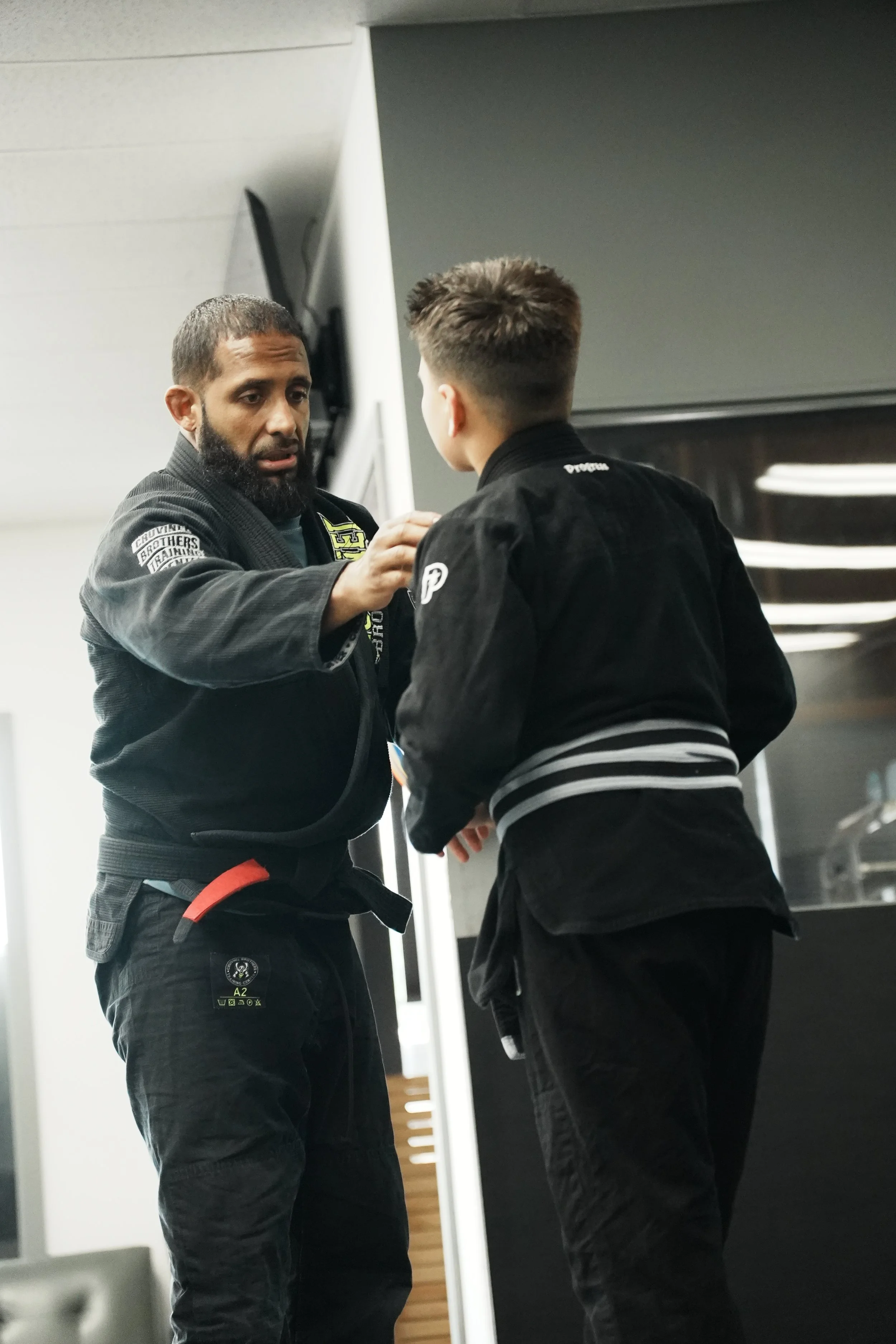Two martial artists, one instructor and one student, wearing black gis and practicing Brazilian Jiu-Jitsu, adjusting the student's collar in a gym.
