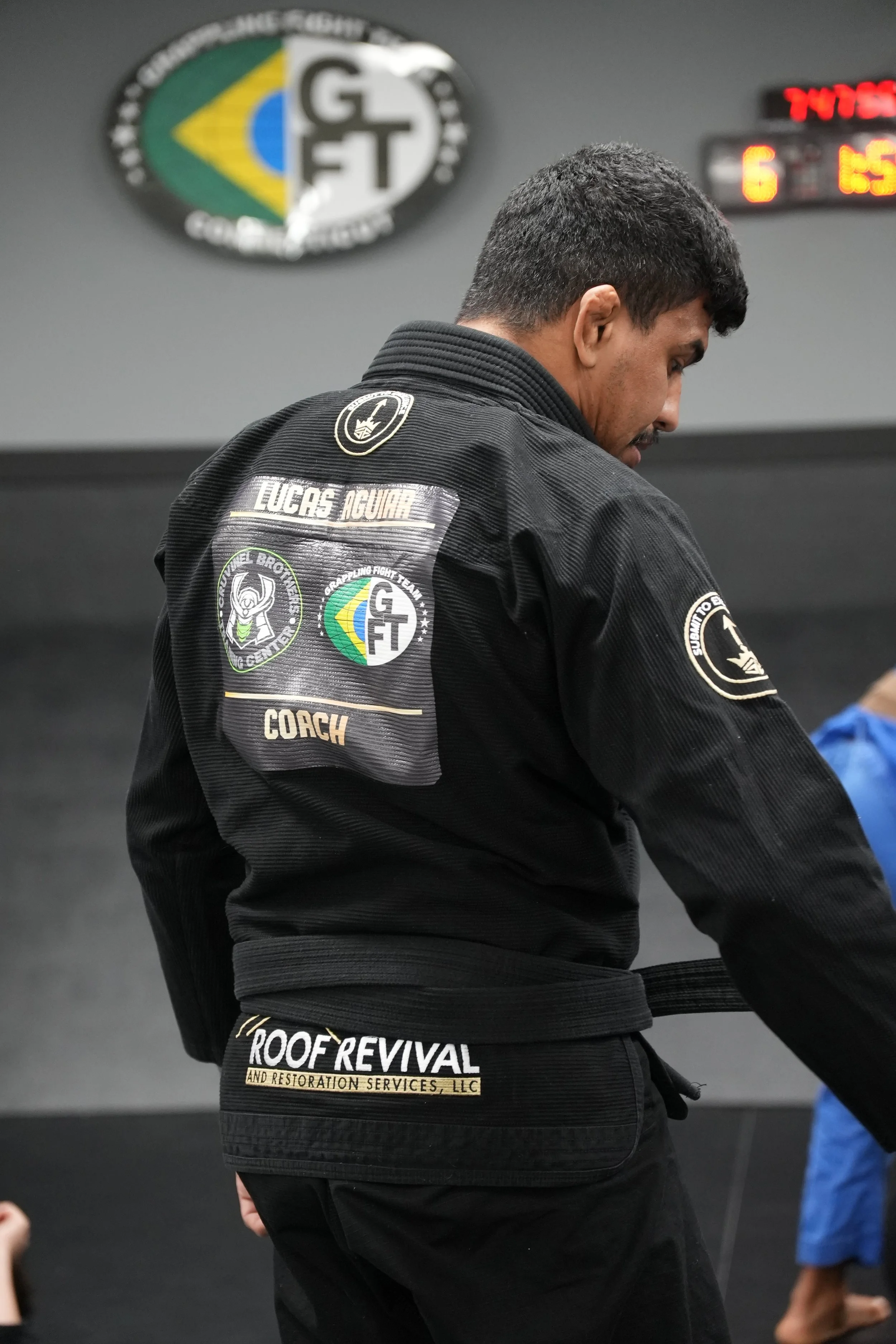 Brazilian Jiu-Jitsu practitioner in black gi with patches, standing in a gym with a Rio de Janeiro logo on the wall, and a scoreboard in the background.