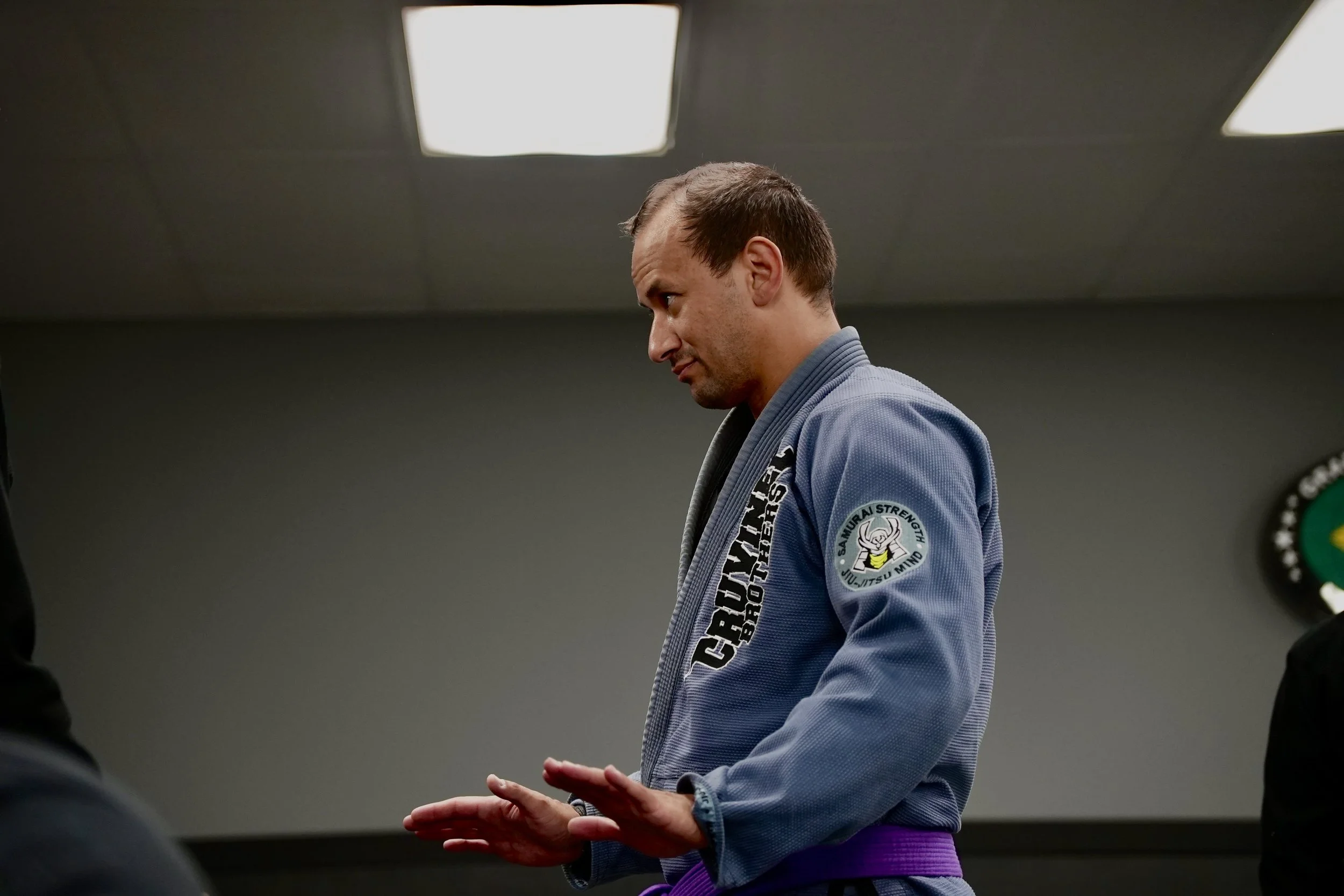 A man in a Brazilian Jiu-Jitsu gi with purple belt standing in a martial arts dojo. He appears to be focused, with his hands positioned as if performing a technique. The background has a gray wall and ceiling lights.