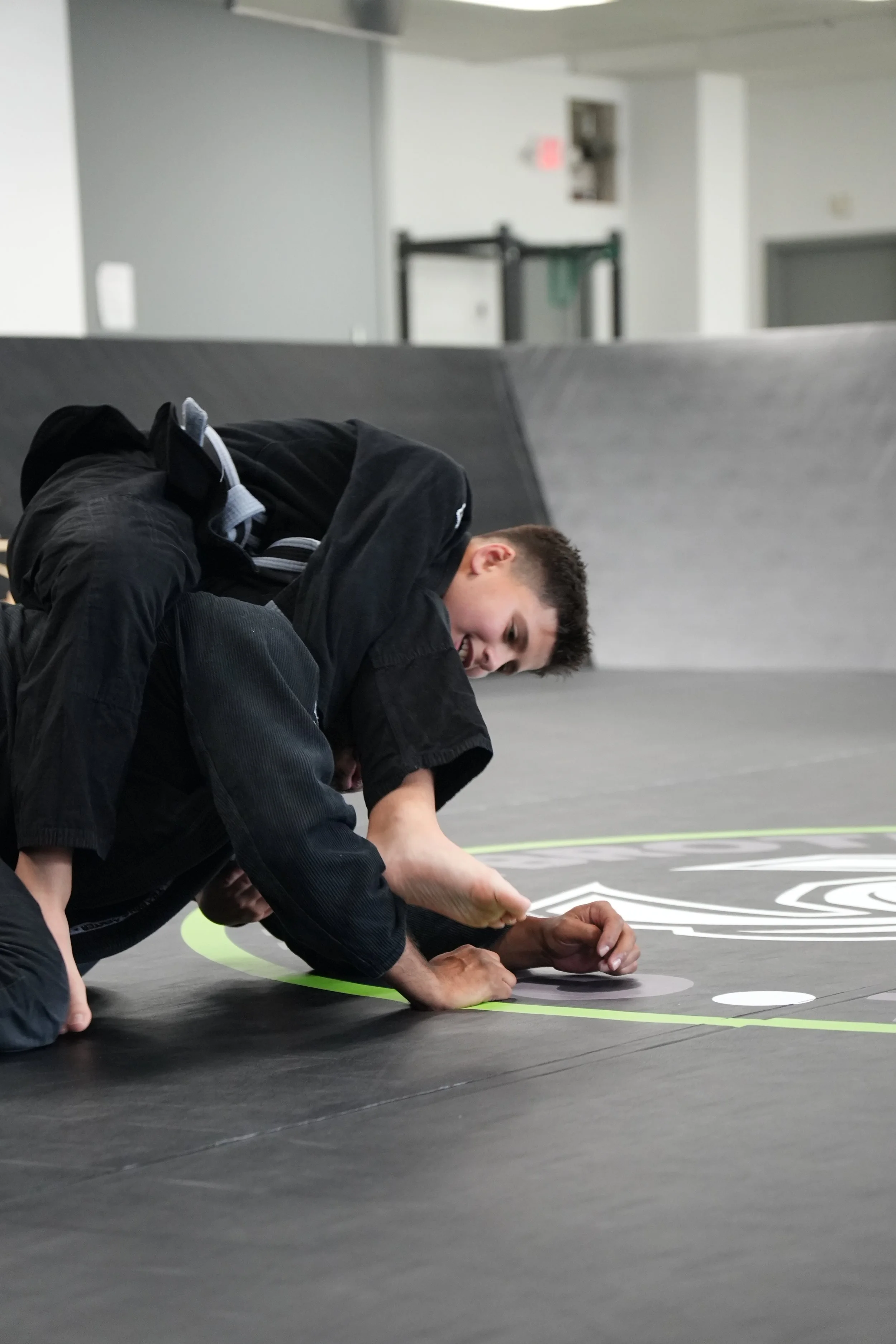 Two martial artists in black uniforms training on a gray mat with white and green markings inside a gym or dojo, with a smiling young man's face visible.