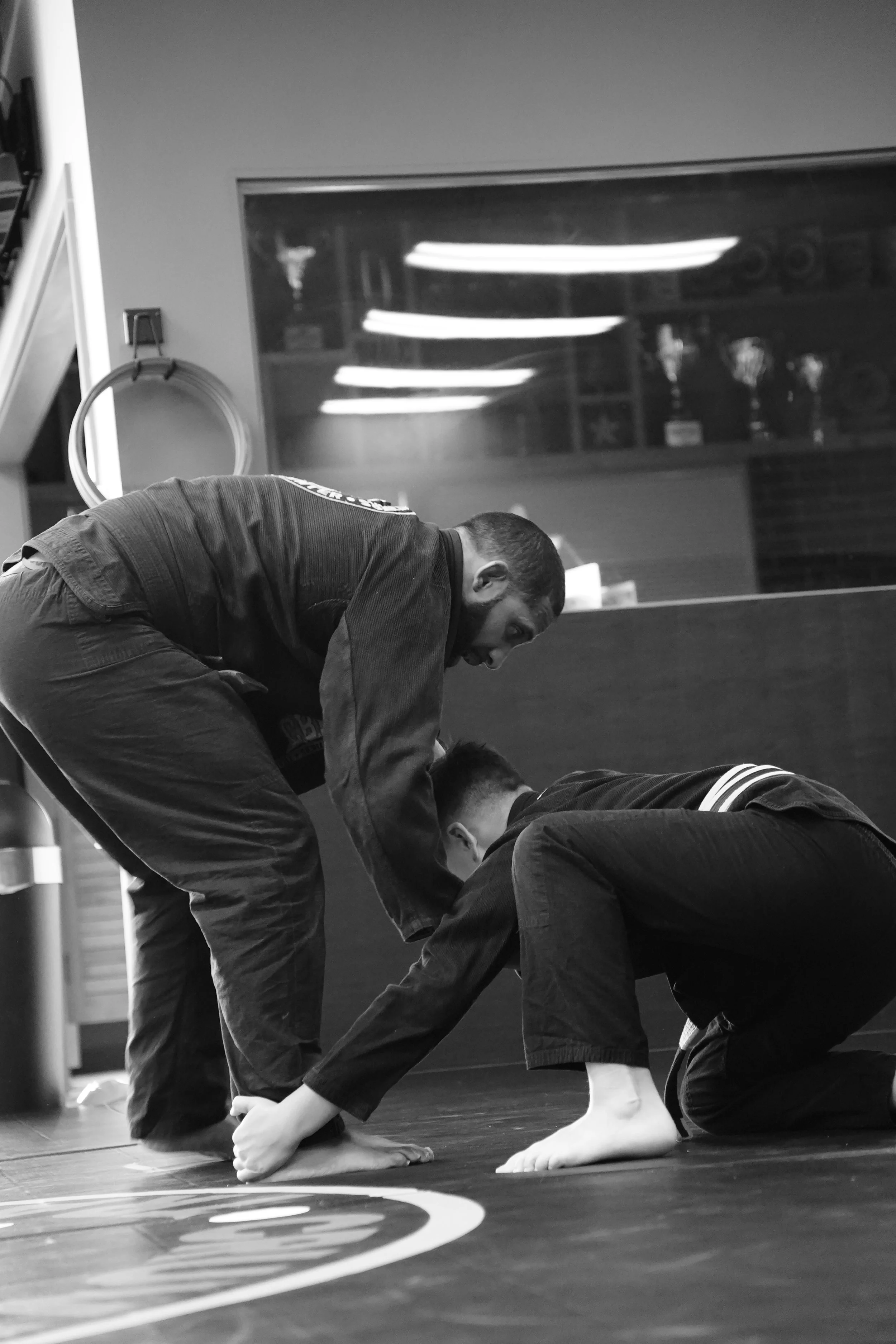 Two individuals practicing martial arts on a mat, one assisting the other. In a practice studio with fluorescent lighting and a window reflecting the room.