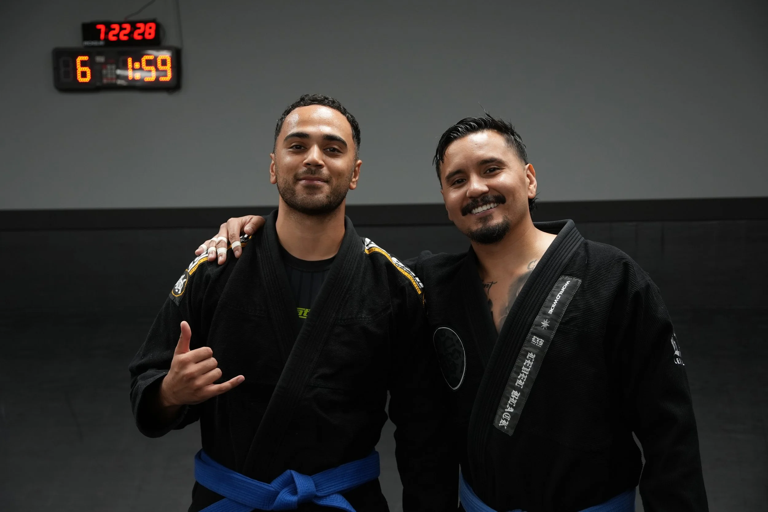 Two men in Brazilian Jiu-Jitsu uniforms smiling and standing together in a gym with a digital clock on the wall showing the time and some numbers.