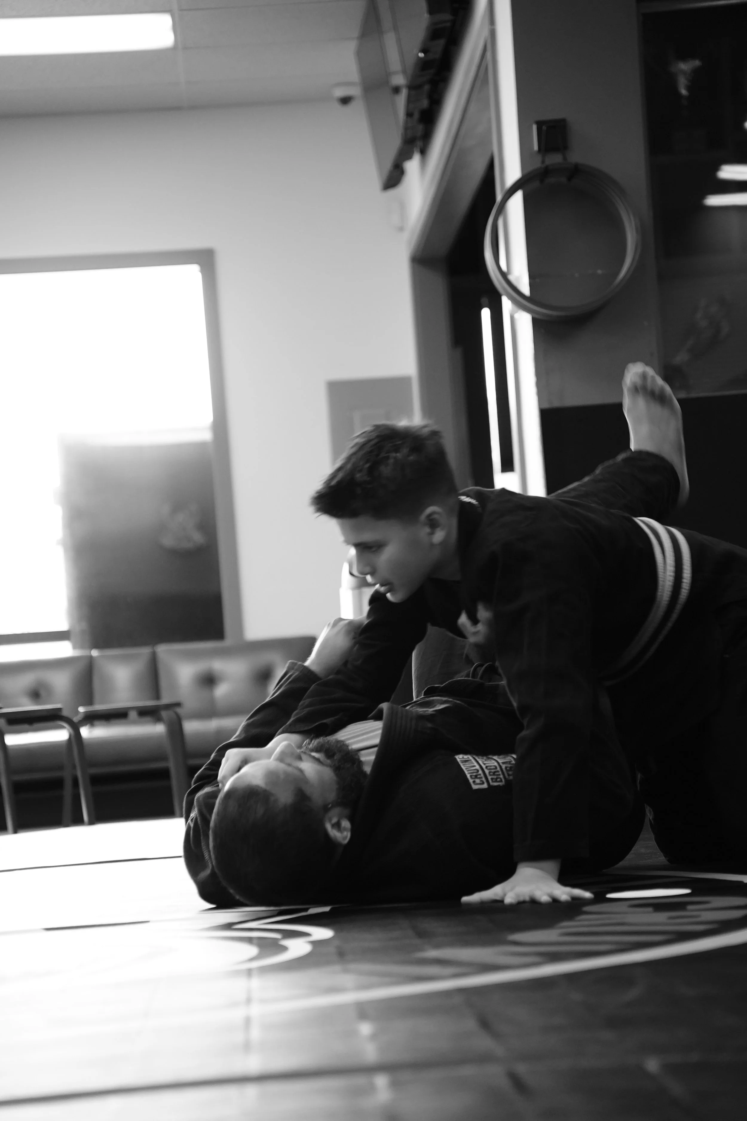 Two young men practicing Brazilian Jiu-Jitsu on a mat in a martial arts school. One is on the ground, and the other is applying a submission hold.