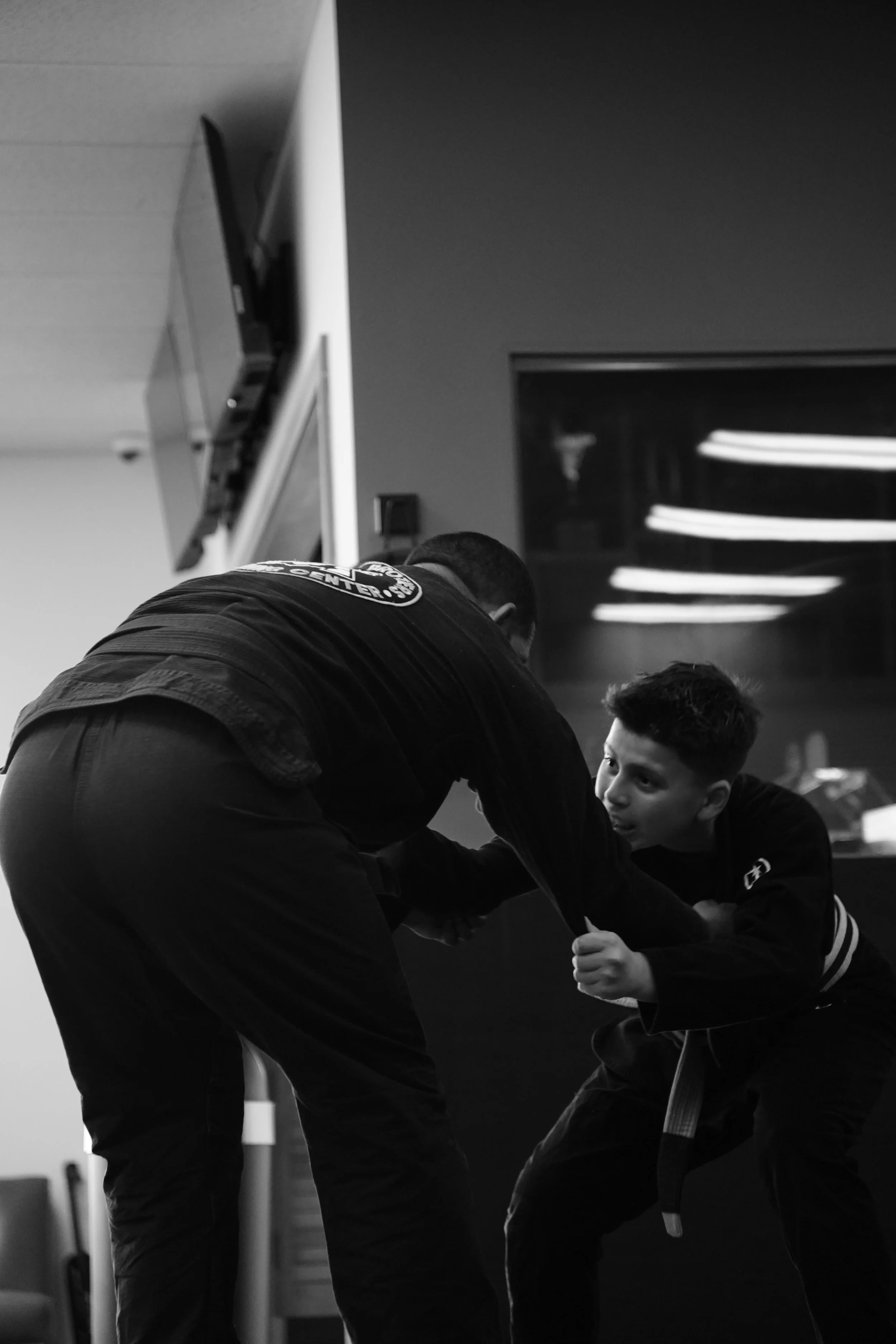 A man and a child practicing martial arts together indoors, with the man bending over and the child gripping his arm in a training stance.
