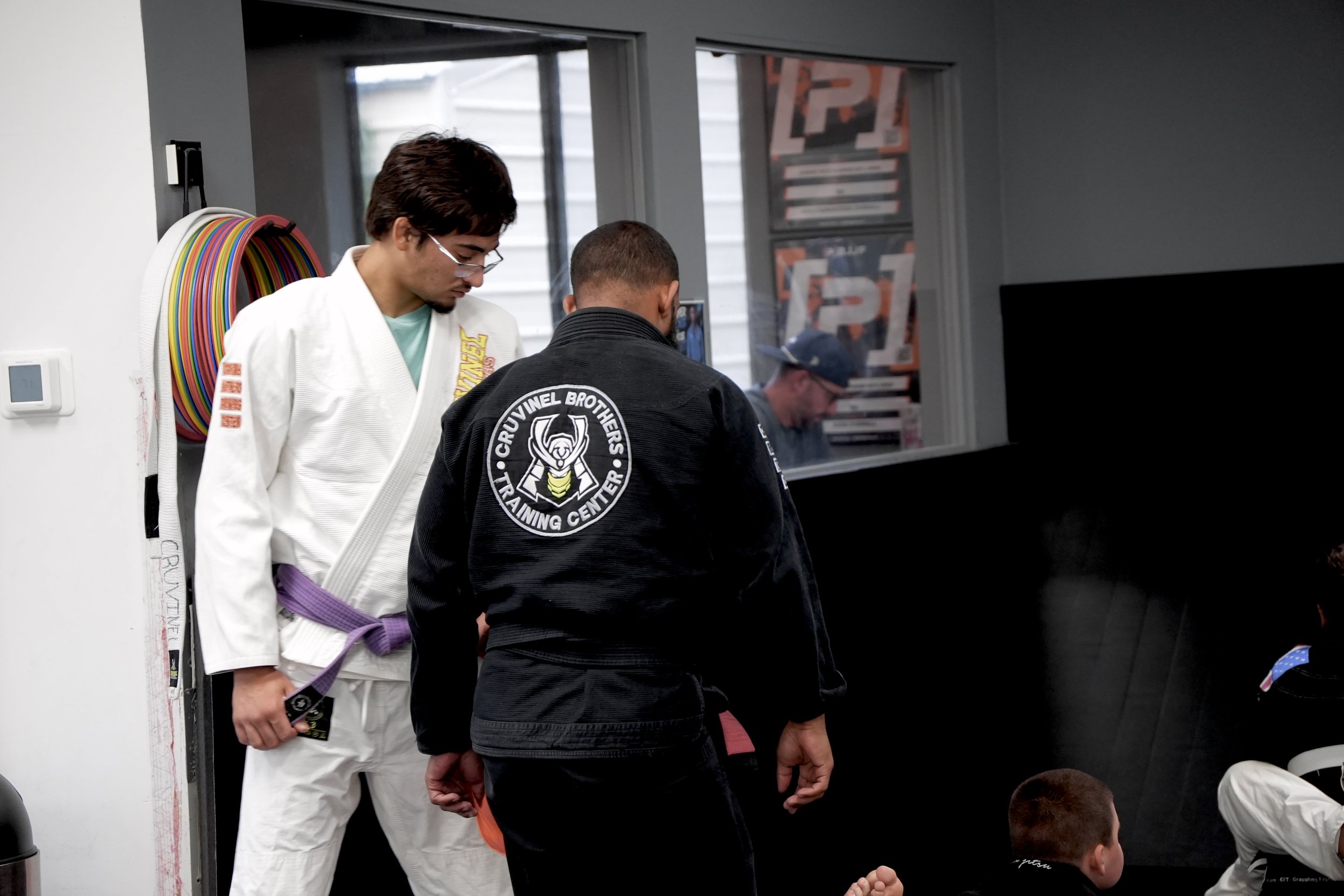 Two men, one in a white jiu-jitsu gi with a purple belt and the other in a black gi with a patch that says 'Cruvinel Brothers Training Center,' stand close to each other in a martial arts training facility. Other children in martial arts gis are visi