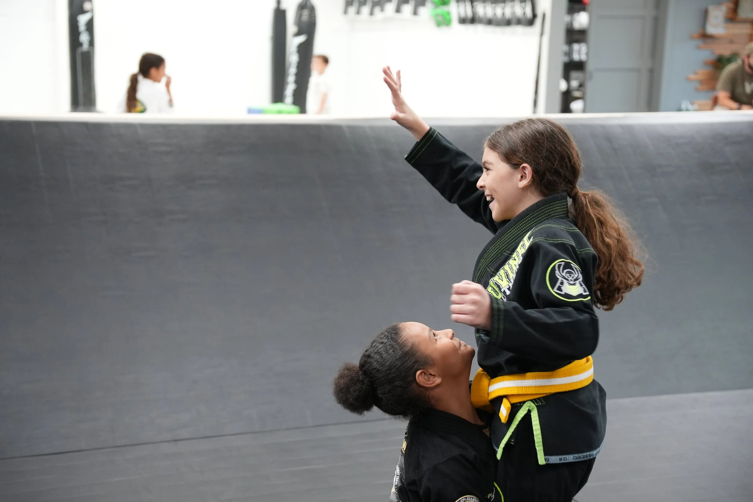 A female martial arts instructor holding a young girl in her arms during a training session in a gym. The girl is reaching out with her right hand in a punching or striking pose, while the instructor supports her from below, both smiling. The backgro