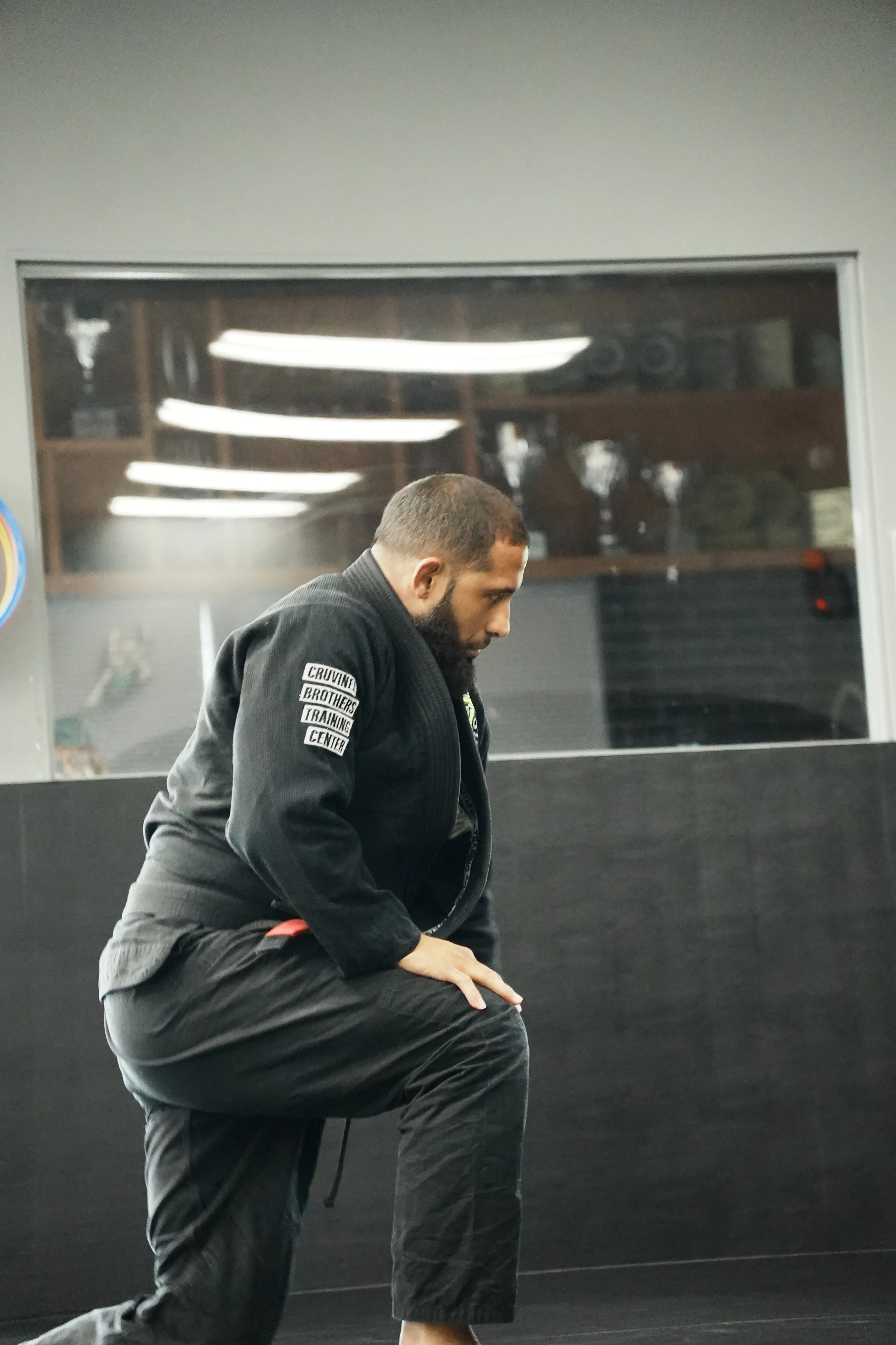 A man in a black gi practicing martial arts, in a dojo or training center, with a black wall and windows behind him.