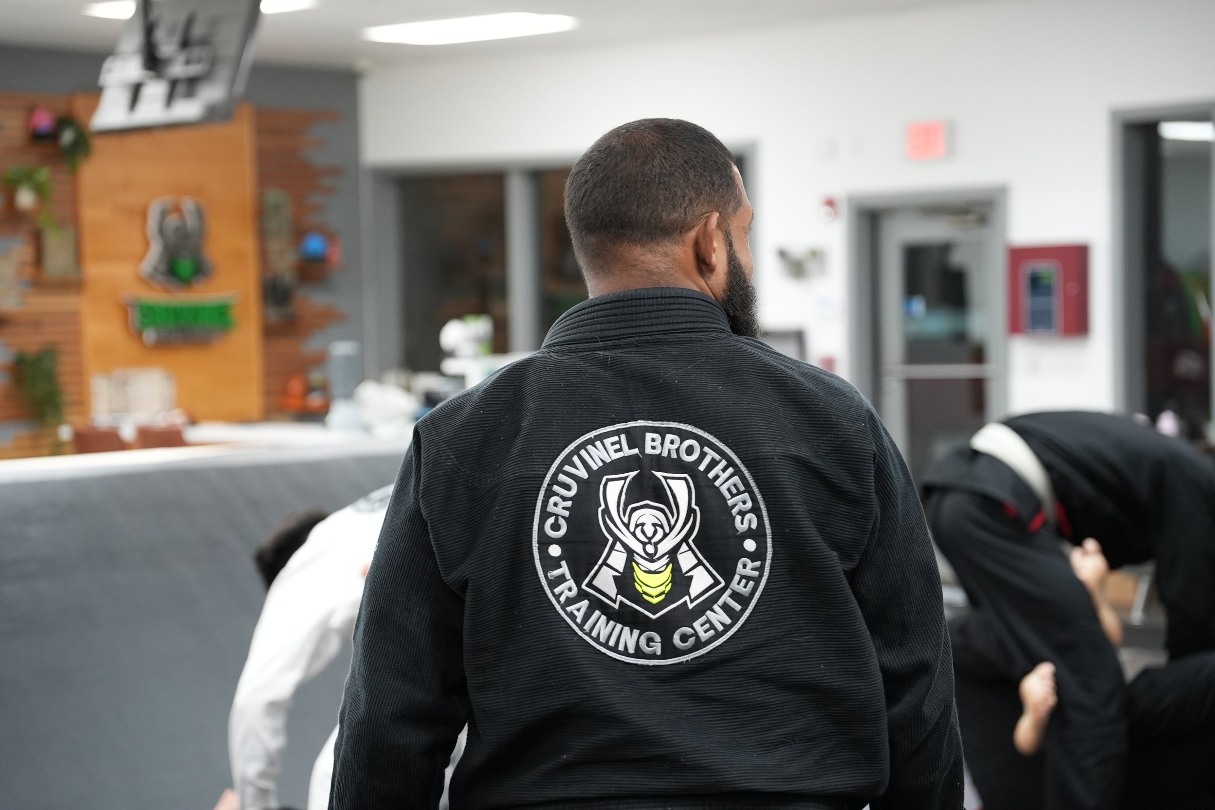 Back view of a man wearing a black martial arts gi with a crest that reads "Crucible Brothers Tactical Training Center" on the back, standing inside a martial arts training facility.
