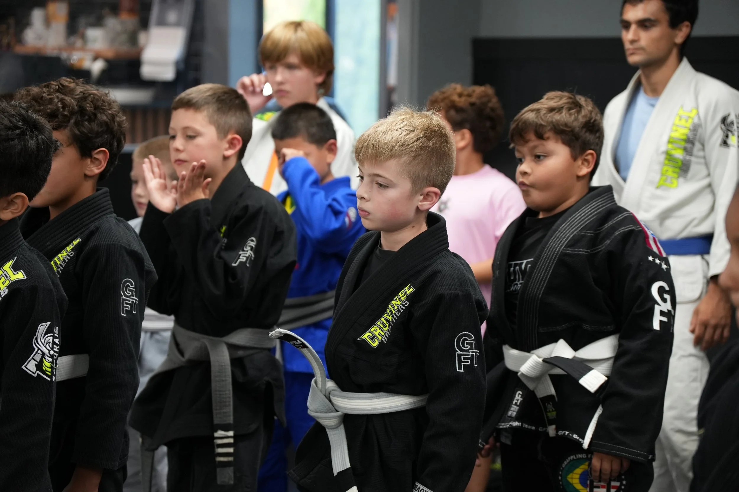 Group of children in martial arts uniforms standing with hands raised in a stance. An instructor in a white gi with yellow lettering stands nearby.